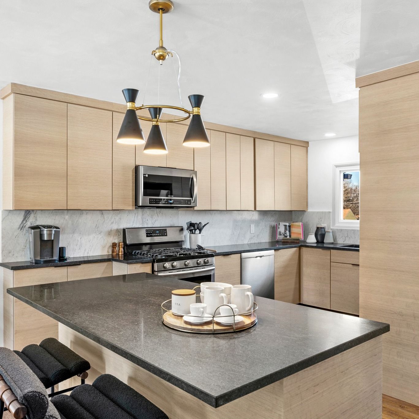 Modern kitchen with light wood cabinets, dark countertops, island with seating, and chandelier.