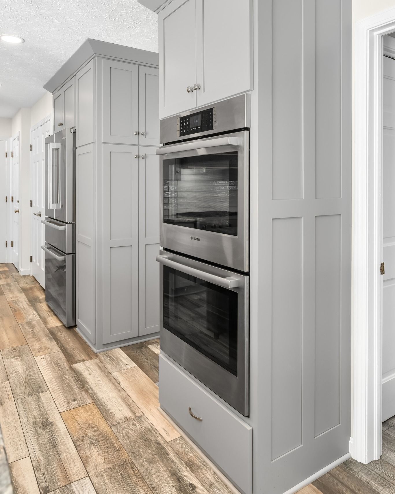 Gray kitchen cabinets with a double oven and refrigerator on wood-look floor.