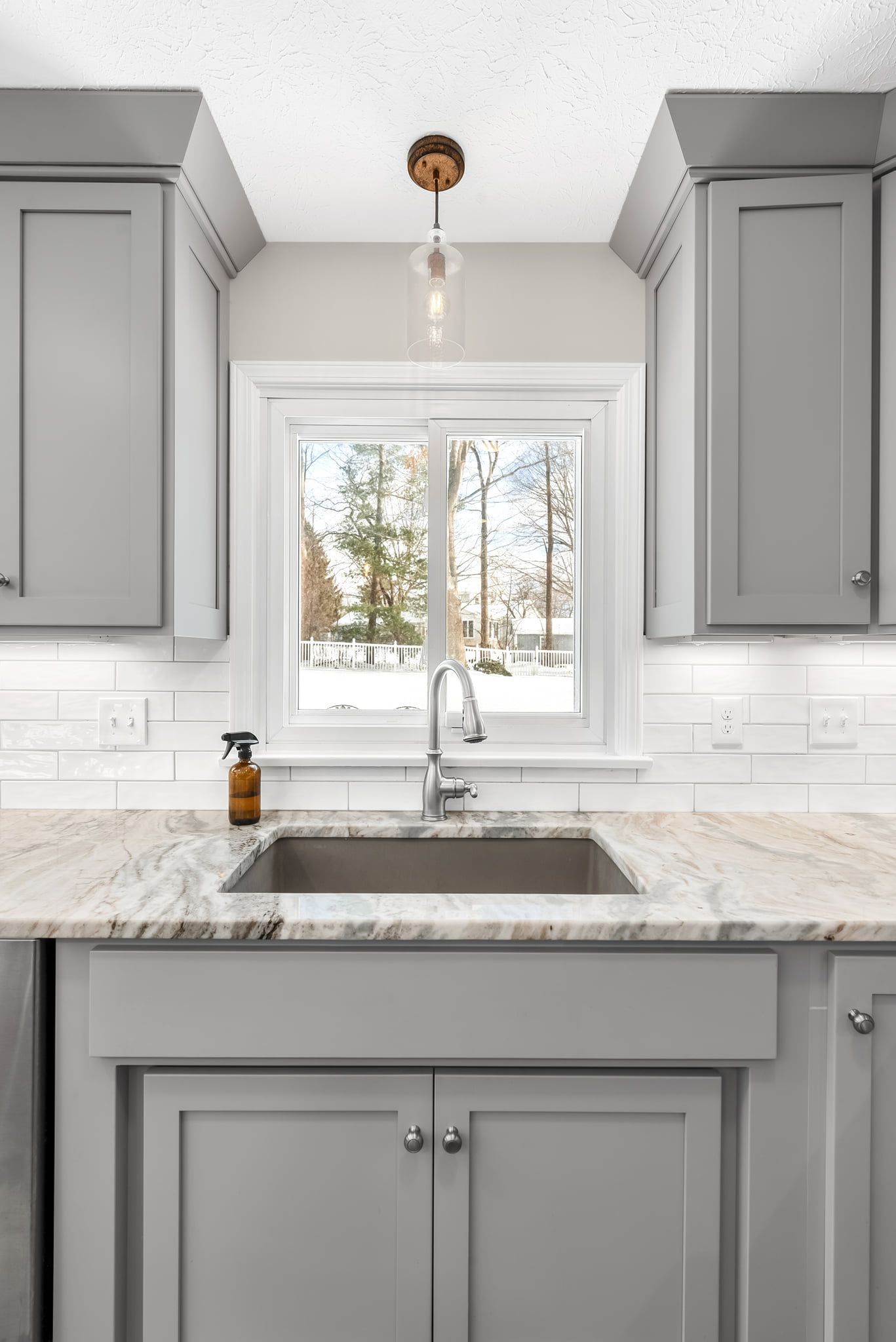 Gray kitchen sink with cabinets, window with trees outside, and pendant light.