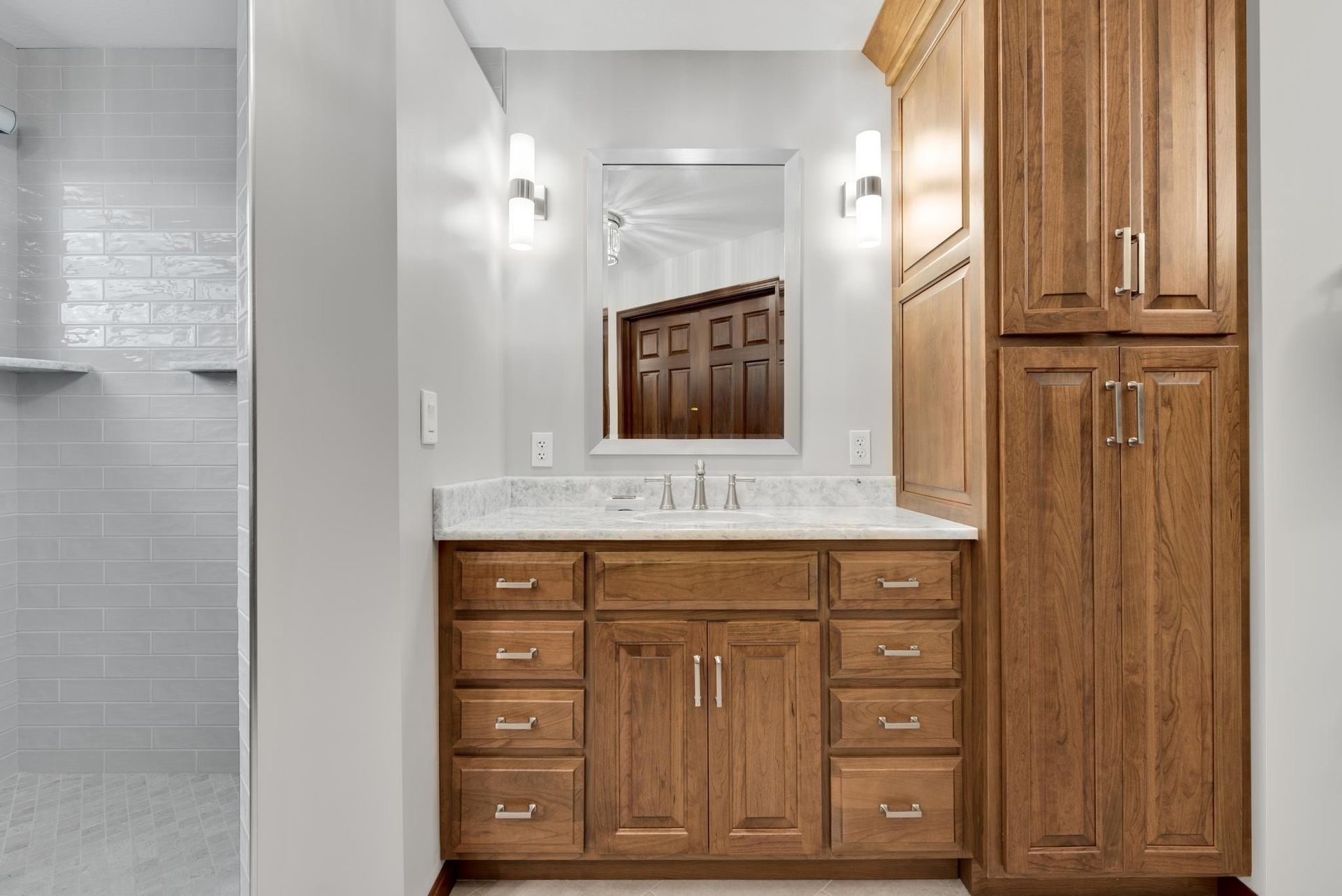 Bathroom with brown wood vanity, tall cabinet, and a tiled shower.