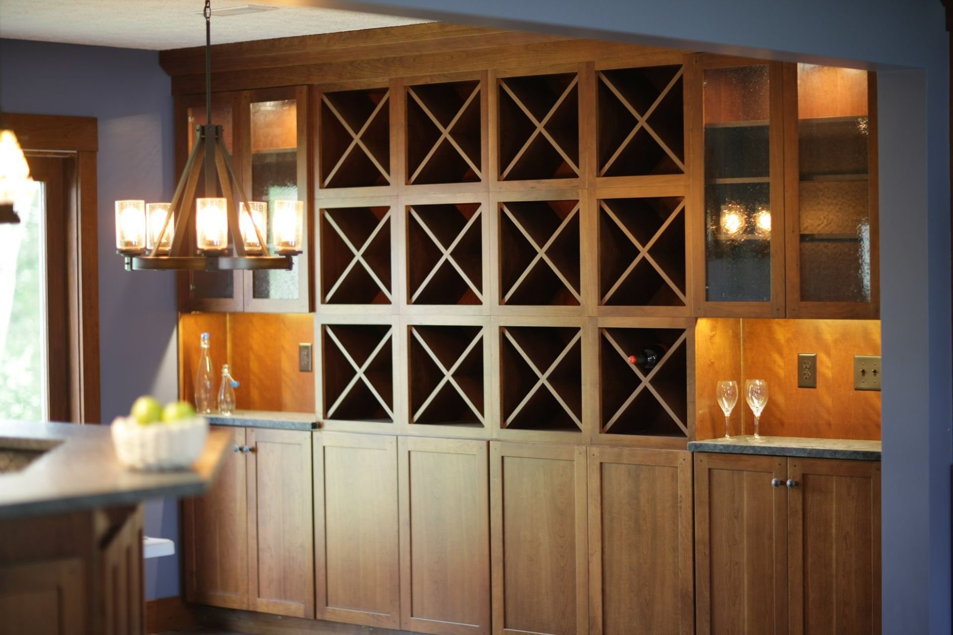 Built-in wooden wine rack with storage cabinets, lit by a chandelier, in a kitchen setting.
