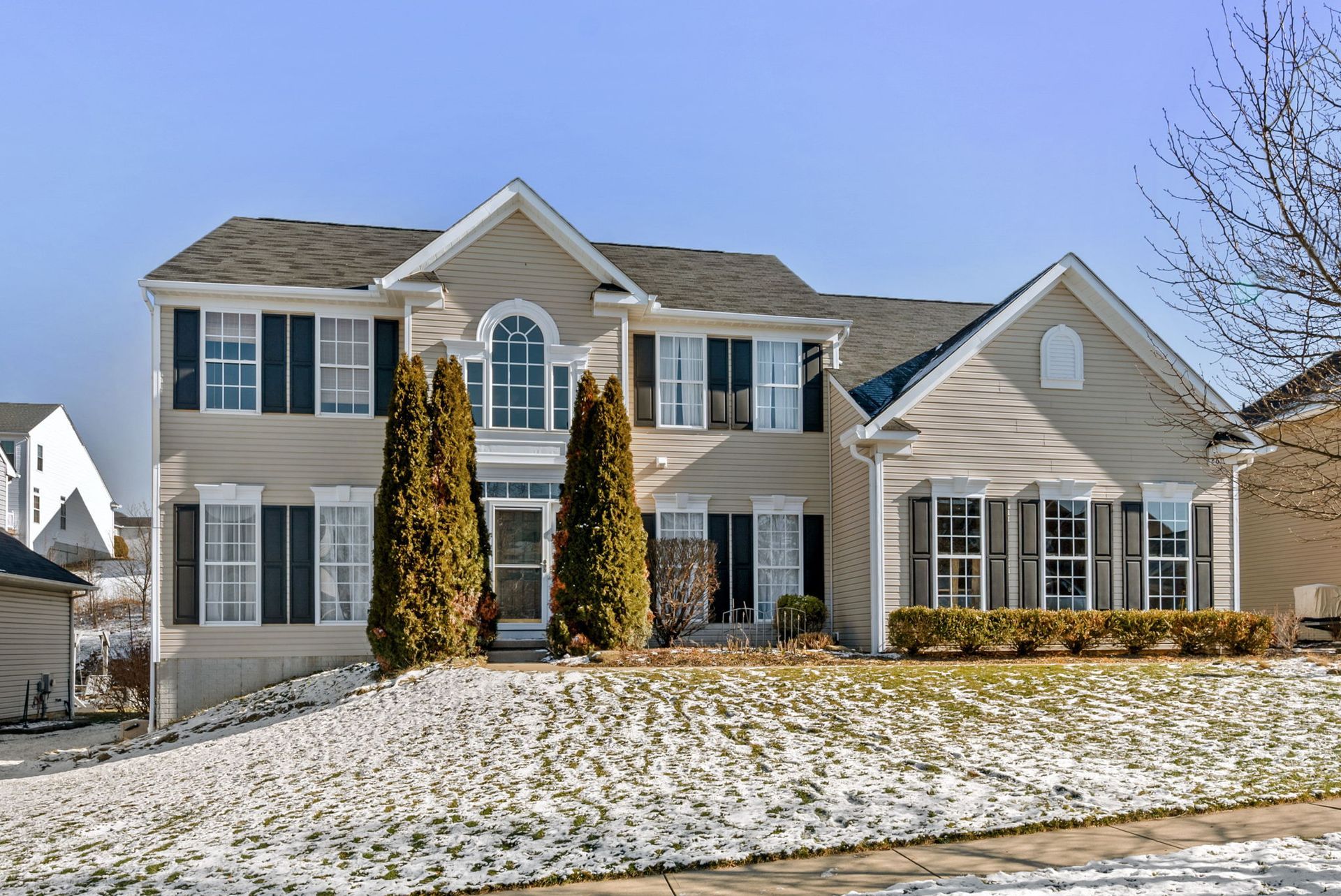 Two-story beige house with black shutters, snow on the lawn, and blue sky.