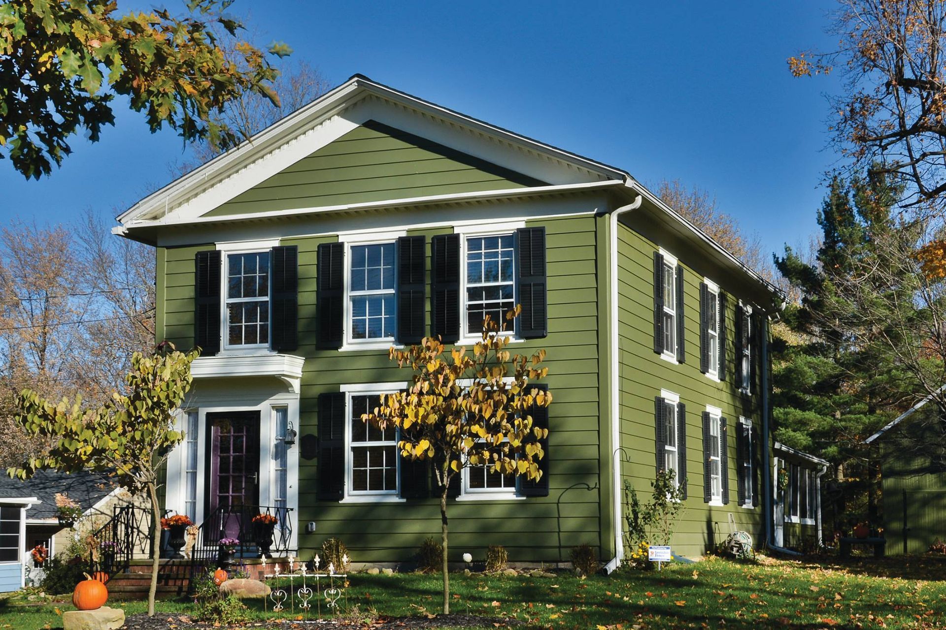 Green two-story house with black shutters, white trim, and a small front yard with trees under a blue sky.
