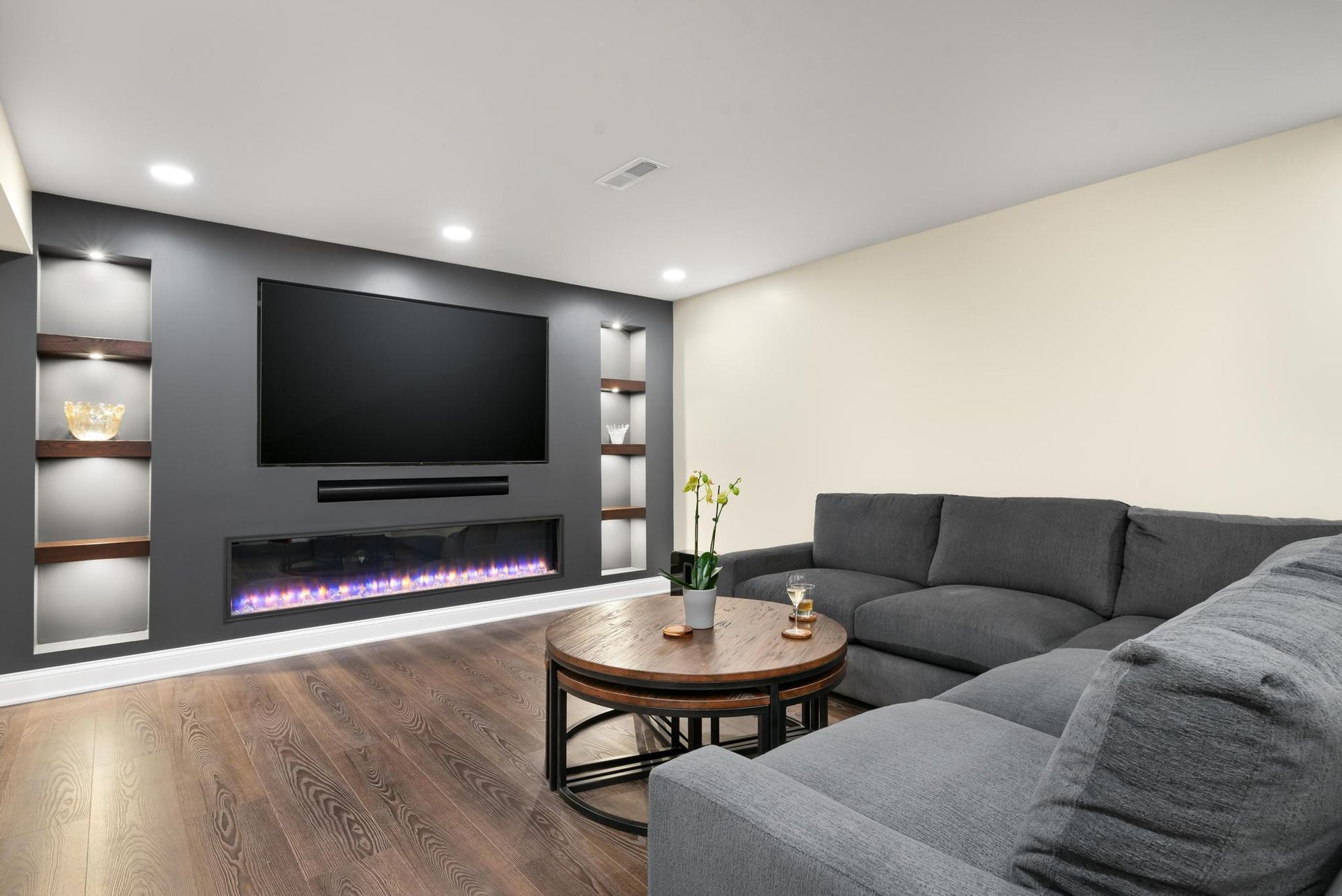 Cozy modern living room with gray sectional sofa, dark accent wall with TV and fireplace, recessed shelving.