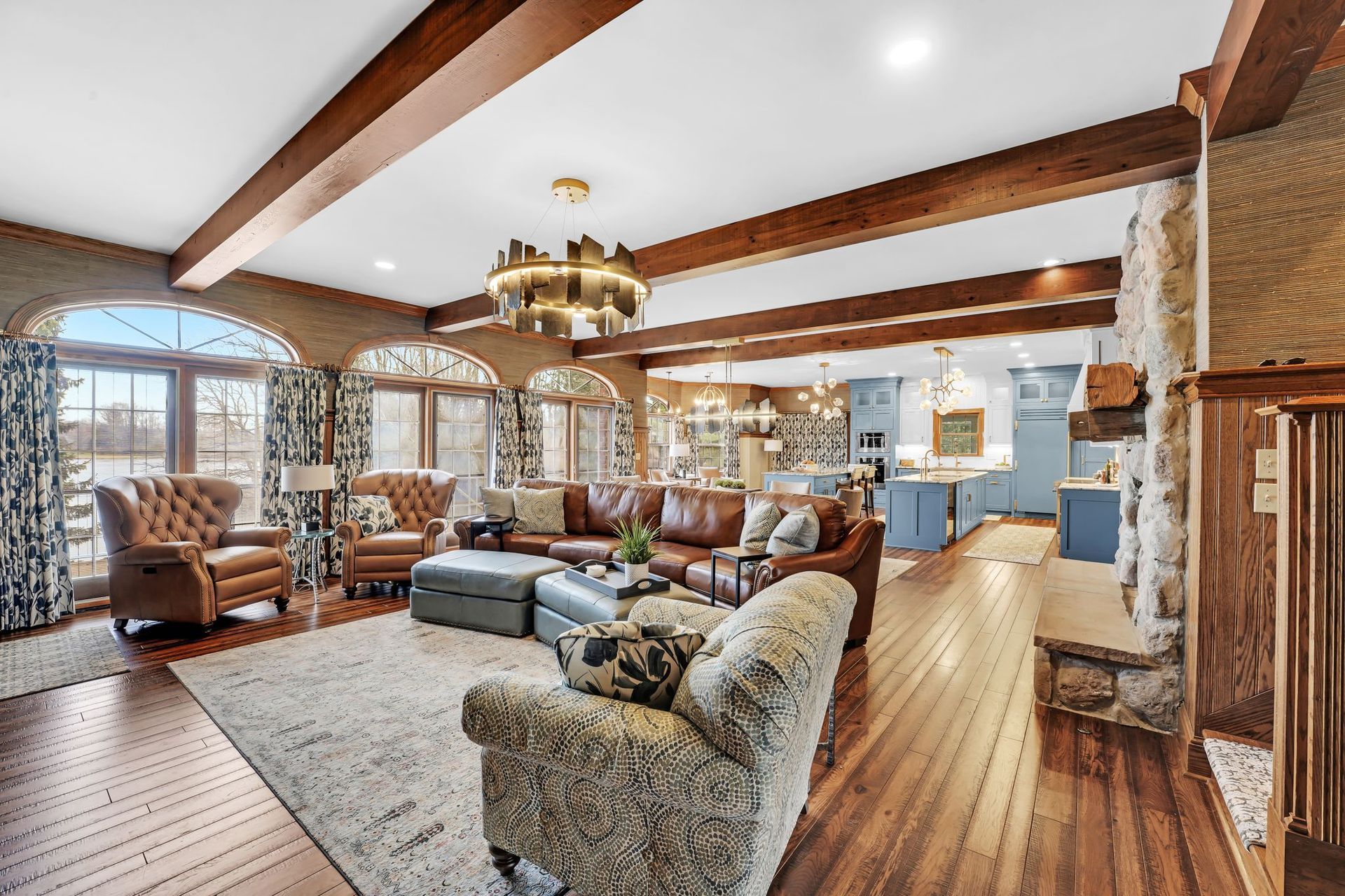 Spacious living room with wood beams, leather sofa, and stone fireplace, leading to a blue kitchen.
