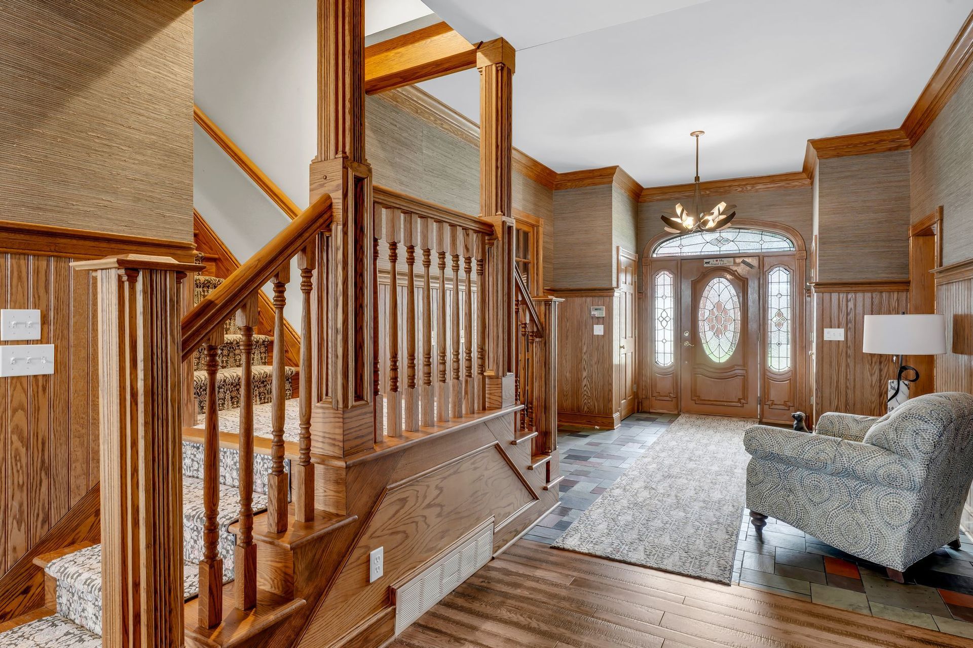 Wooden foyer with a staircase, patterned wallpaper, and a patterned rug near a front door.