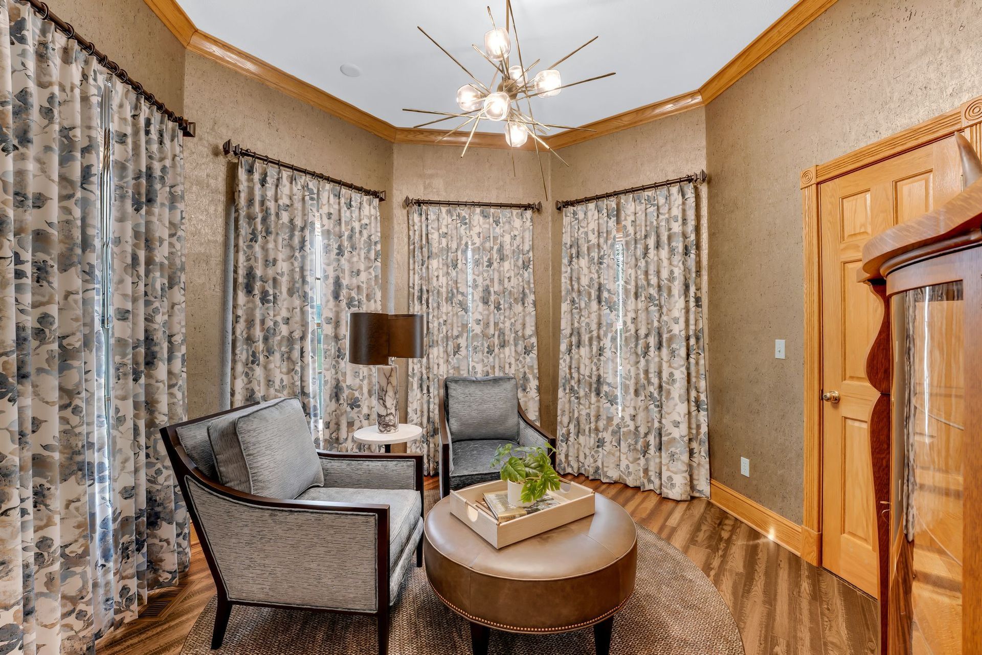 Cozy reading nook with patterned curtains, two armchairs, a round ottoman, and a modern chandelier.