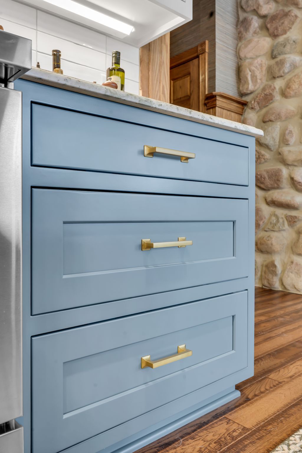 Blue kitchen drawers with gold hardware. Stone wall and wooden floor in the background.