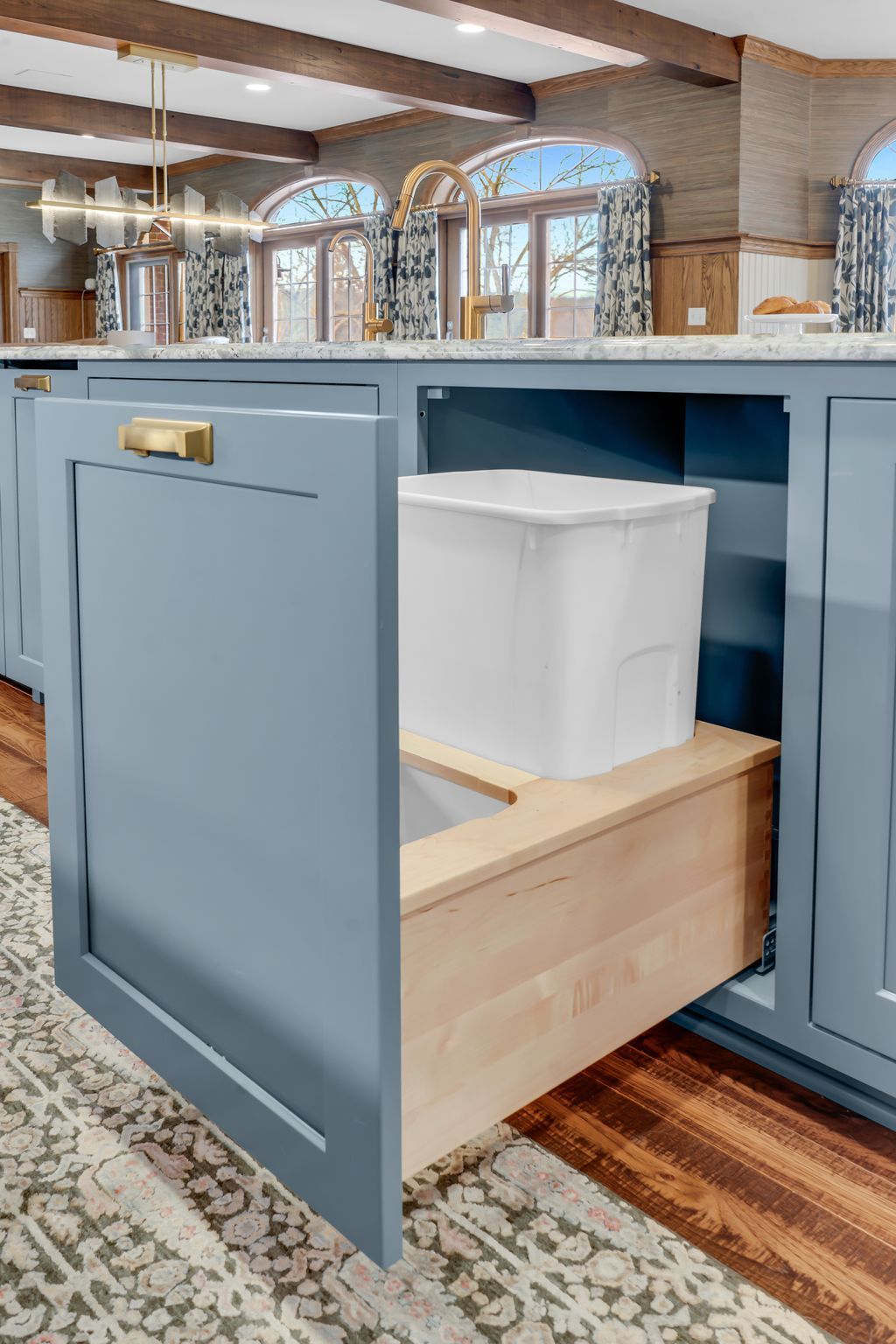 Light blue kitchen cabinet with pull-out trash bin and gold handle.