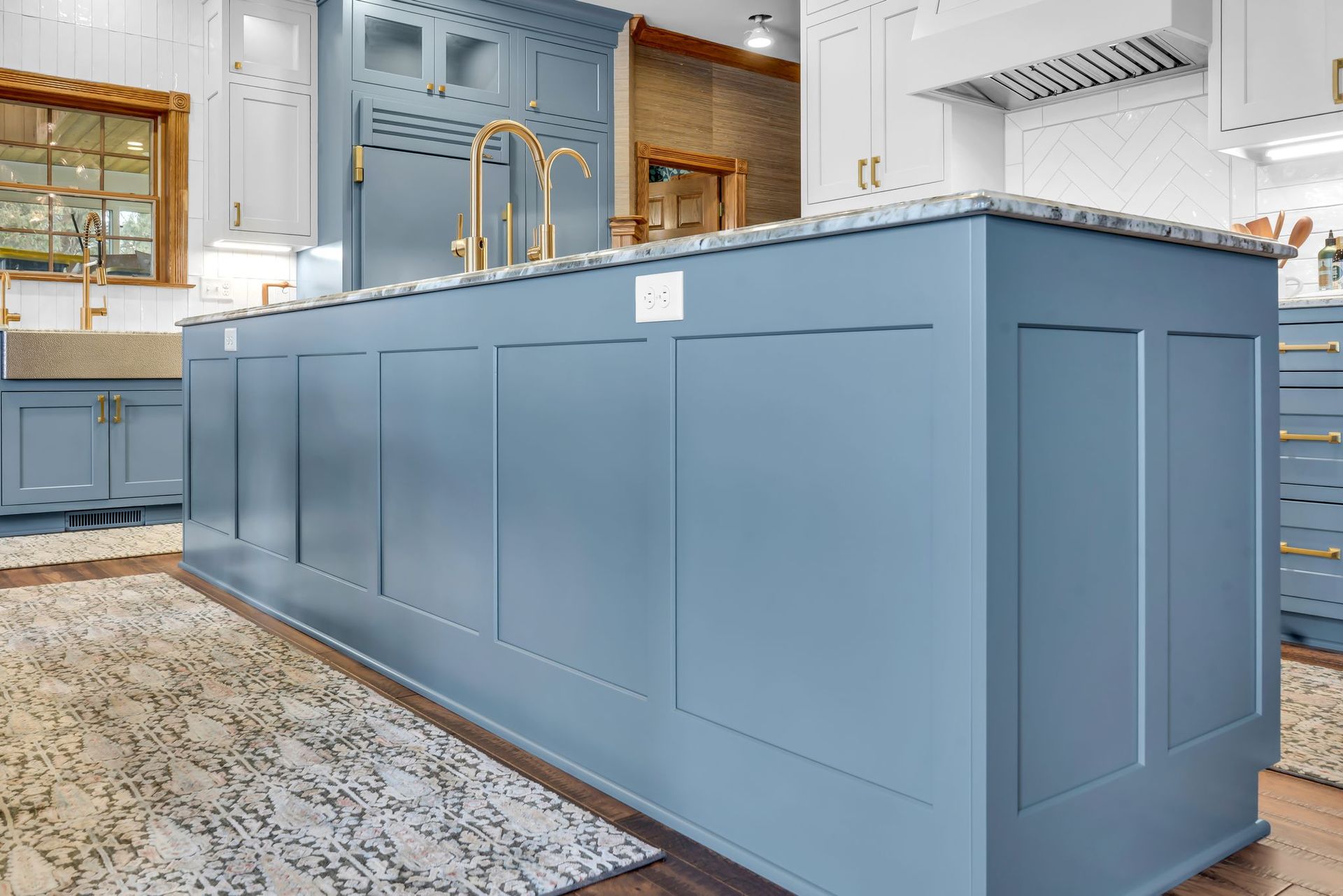 Blue kitchen island with paneling, gold faucet, and a patterned rug.