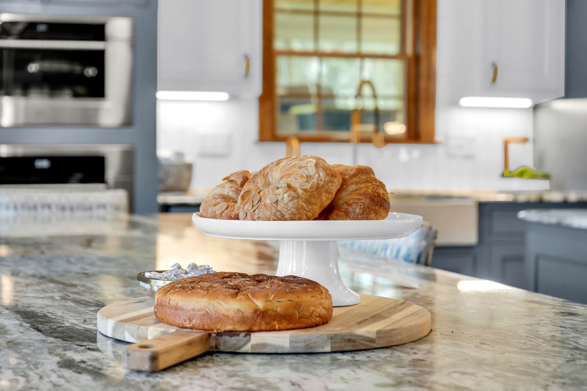 Bread display on a kitchen island: rolls on a cake stand and a loaf on a wood cutting board.