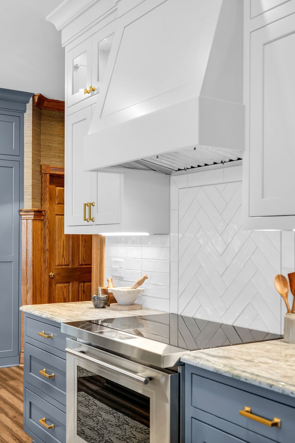 White kitchen hood and cabinets over a stove with blue cabinets and gold hardware. Herringbone tile backsplash.