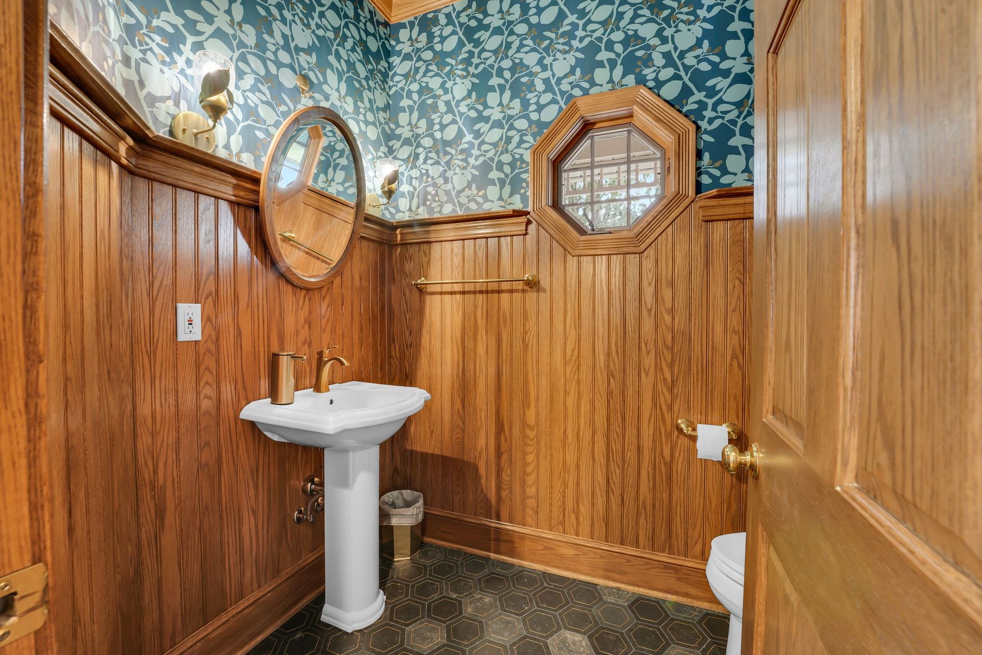Small bathroom with wood paneling, wallpaper, pedestal sink, and octagonal window.