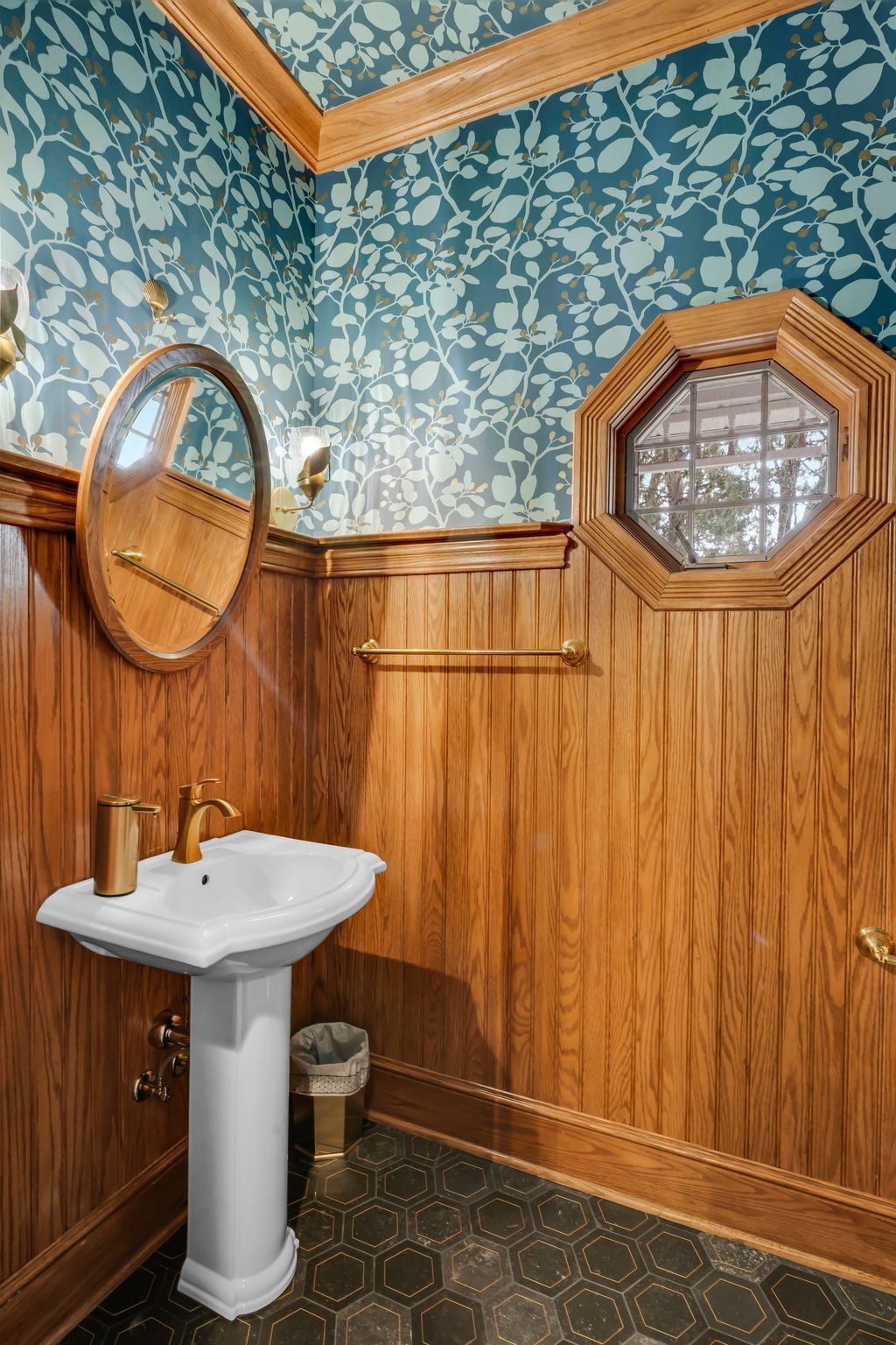 Bathroom with wood paneling, wallpaper, pedestal sink, and octagonal window.