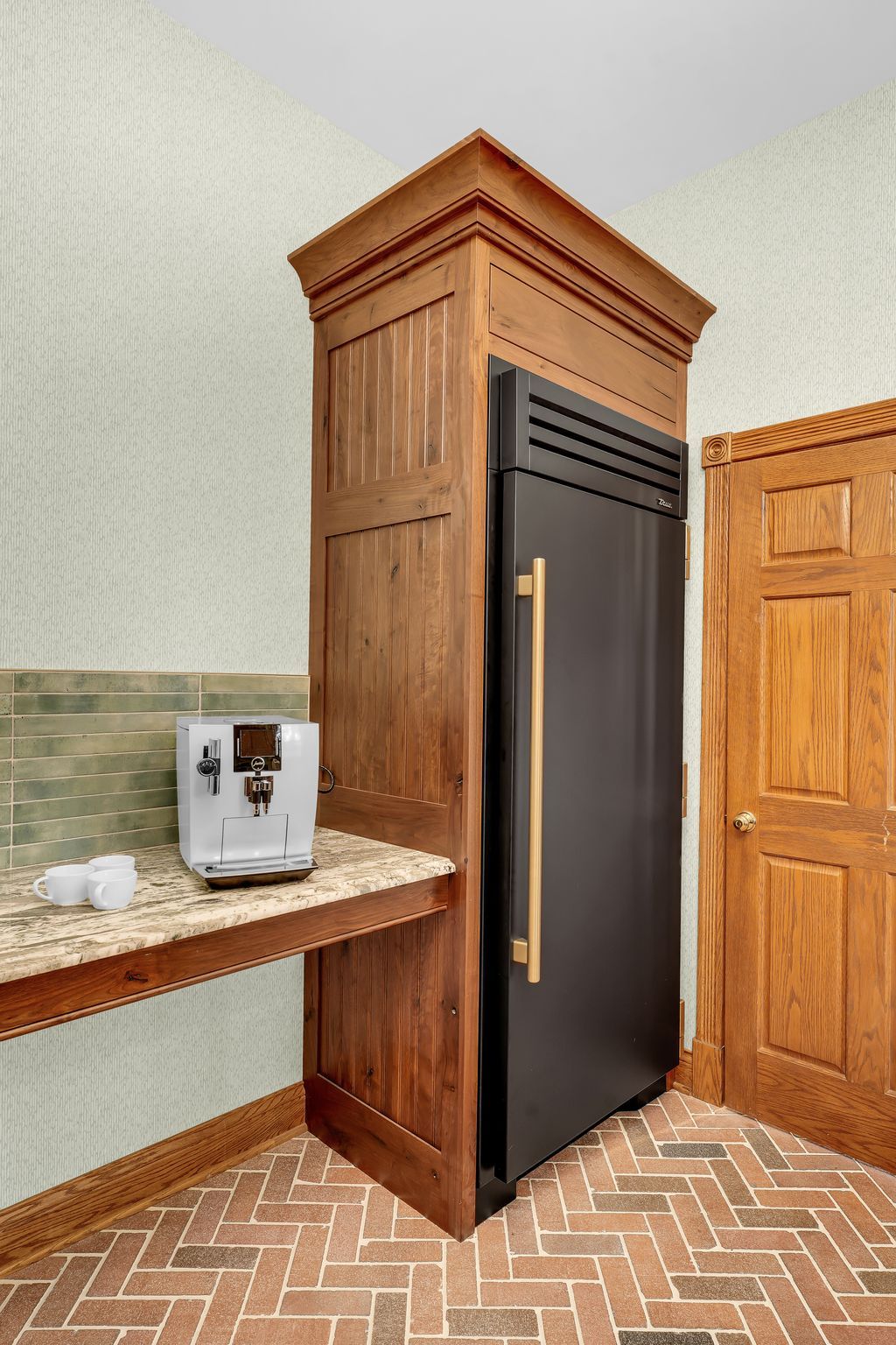 Built-in refrigerator and coffee machine in a wood cabinet with a countertop. Brick floor.