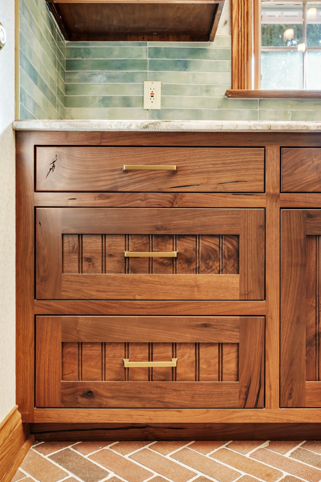 Wooden bathroom vanity with gold handles, brick floor, and turquoise tile backsplash.