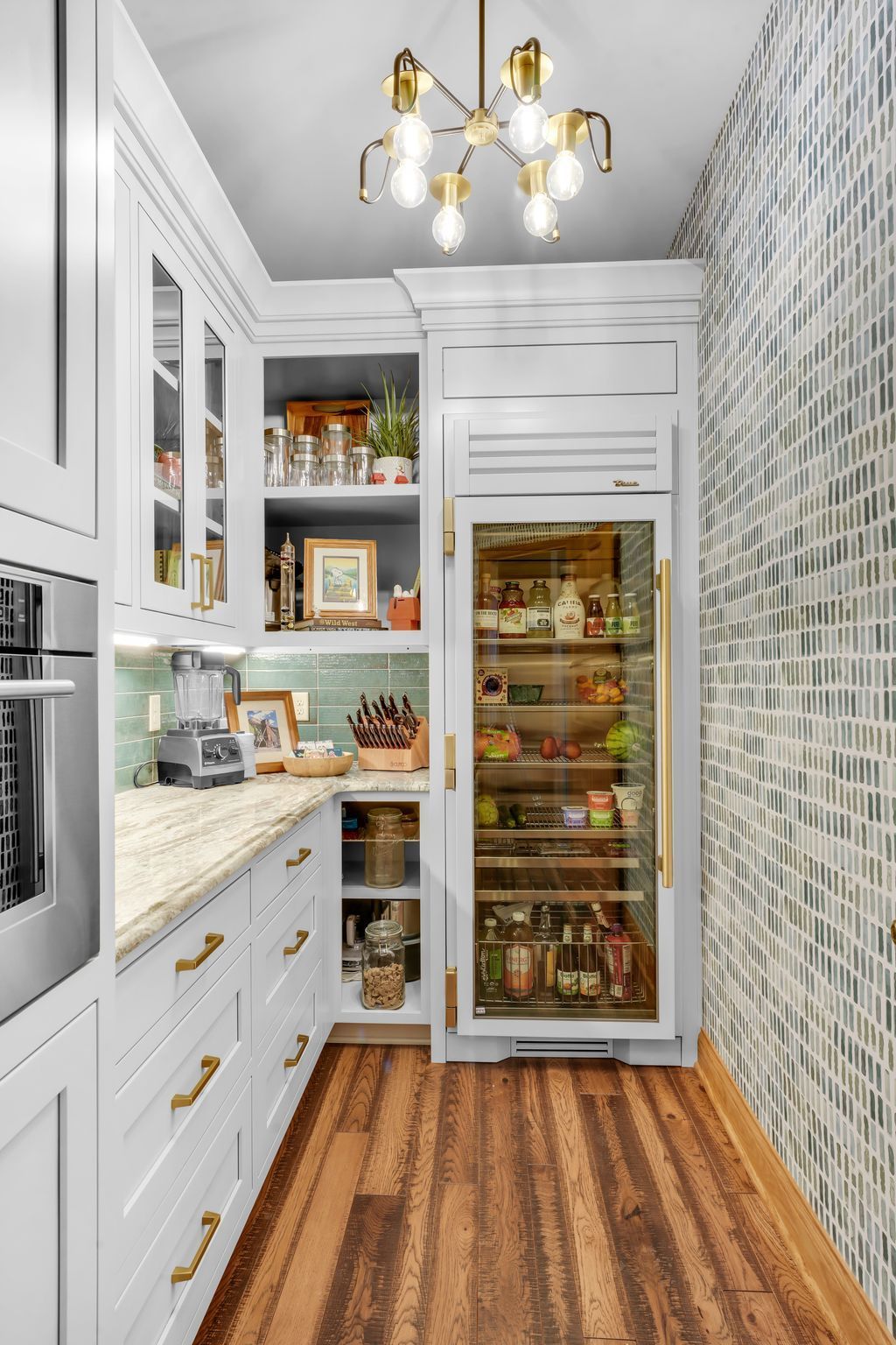 A pantry with white cabinets, gold hardware, built-in refrigerator, and mosaic tiled wall.