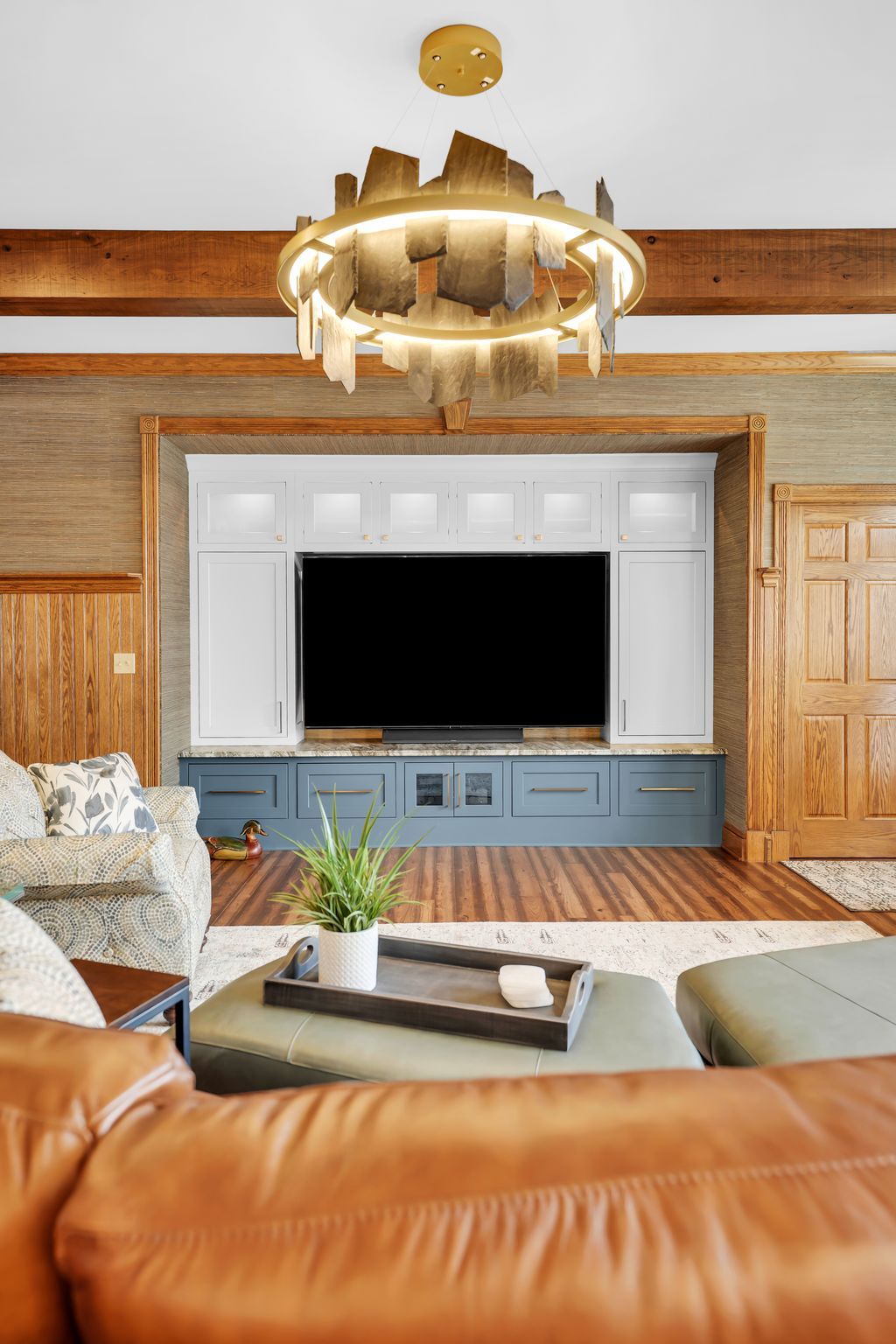 Living room with TV built-in, blue and white cabinets, brown wood beams, gold chandelier, brown leather sectional.