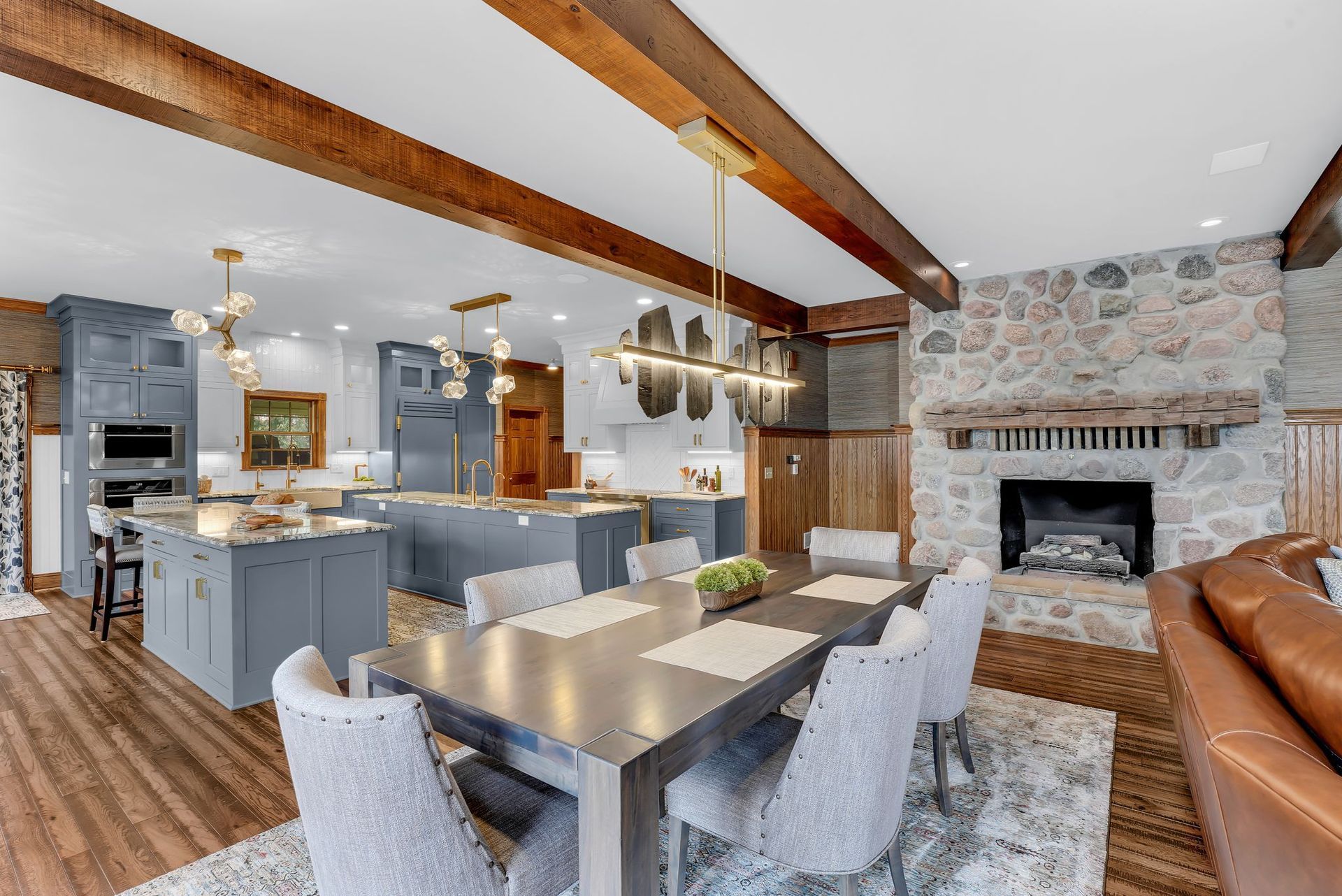 Open-concept kitchen and dining area with gray cabinets, stone fireplace, and wood beams.