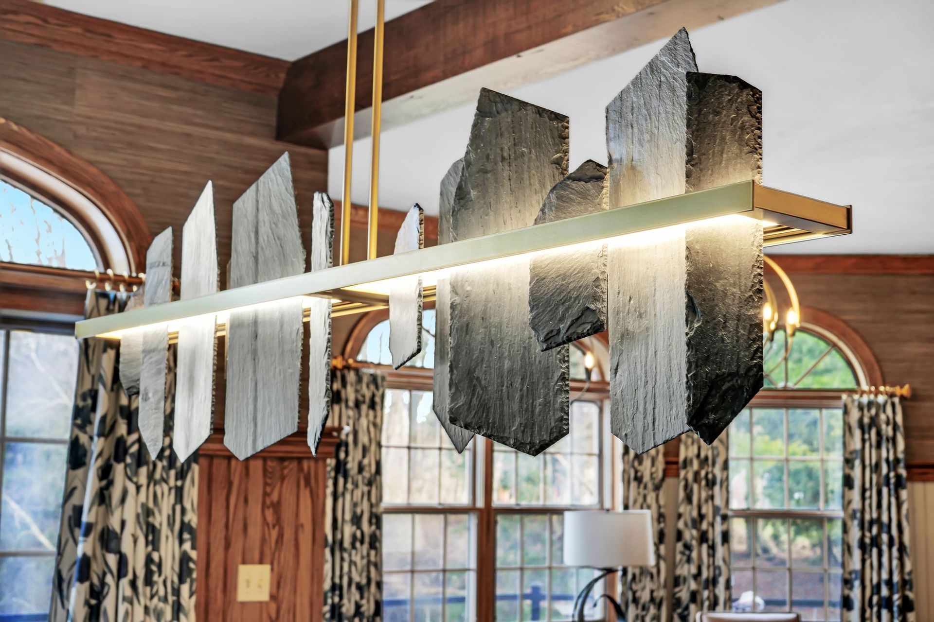 Modern rectangular chandelier with slate-like accents, in a room with wood paneling and arched windows.