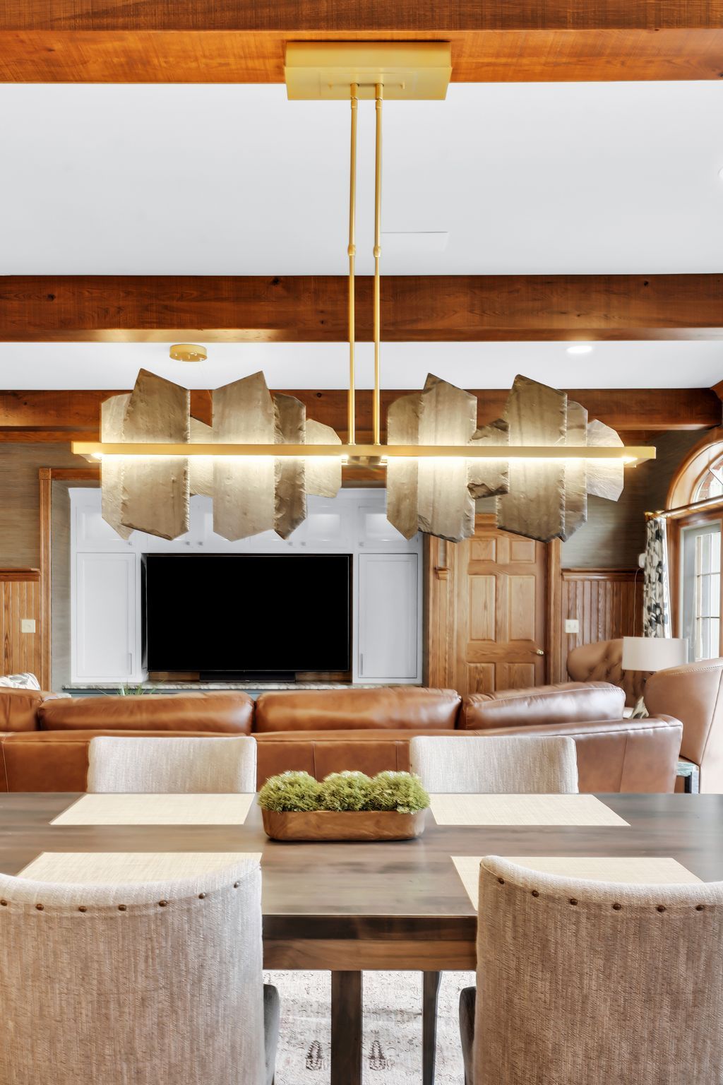 Dining room with modern chandelier over a wooden table and chairs, visible sofa, tv, and wood beams.