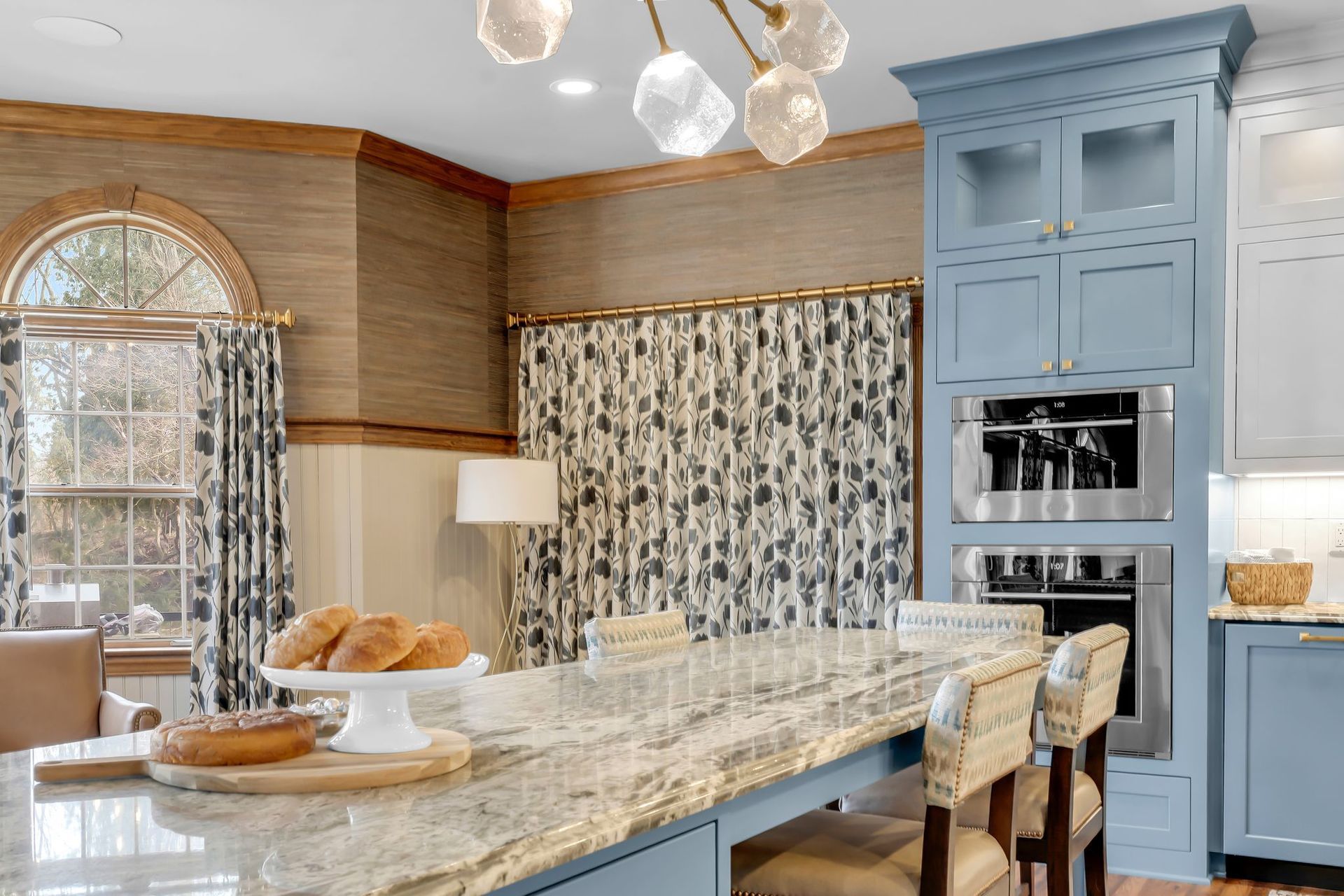 Blue kitchen with island, built-in ovens, and patterned curtains.