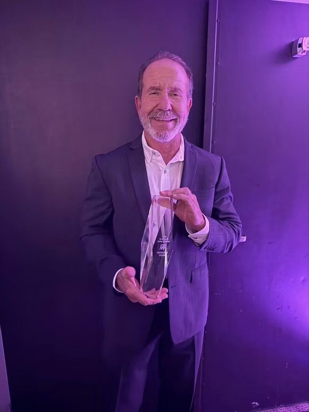 Smiling man in a dark suit holding a glass award under purple lighting against a dark wall