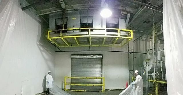 Two people in lab coats working in a warehouse. A loading dock, elevated platform, and industrial equipment are visible.