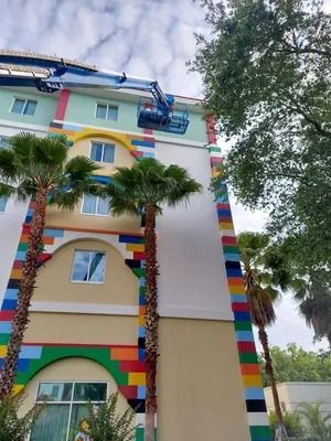 Hotel exterior with colorful brick-like accents, palm trees, and a lift platform with workers near the roof.
