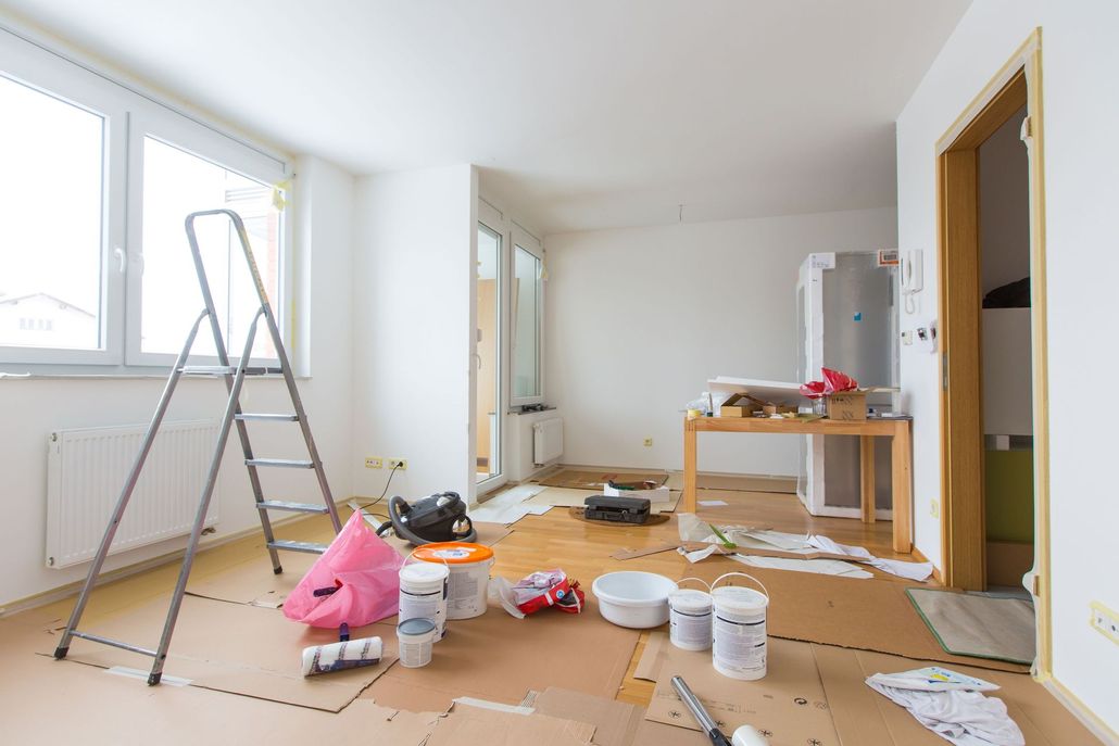 Room in renovation, with ladder, painting supplies, and cardboard on floor.