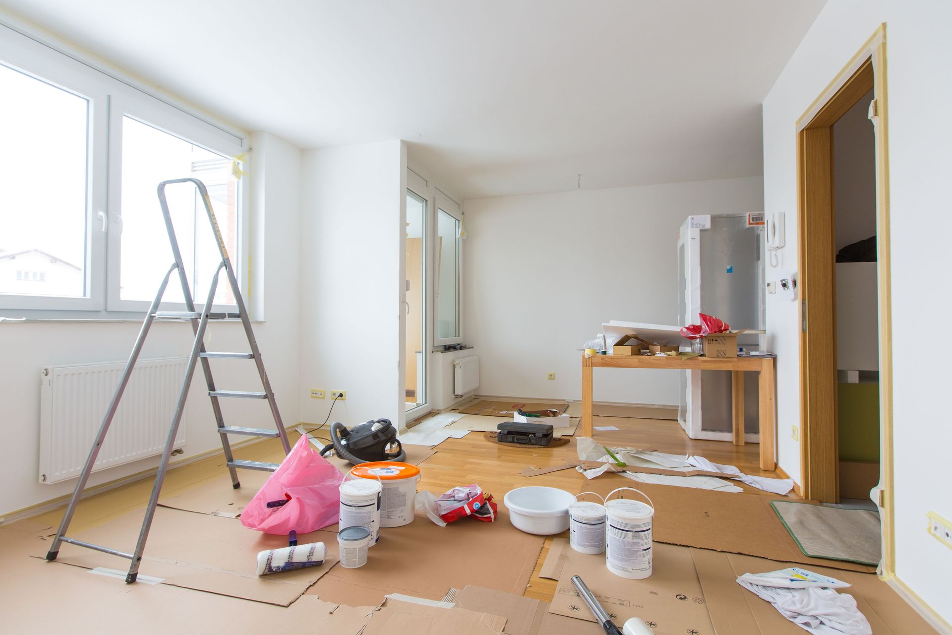 Room in renovation, with ladder, painting supplies, and cardboard on floor.