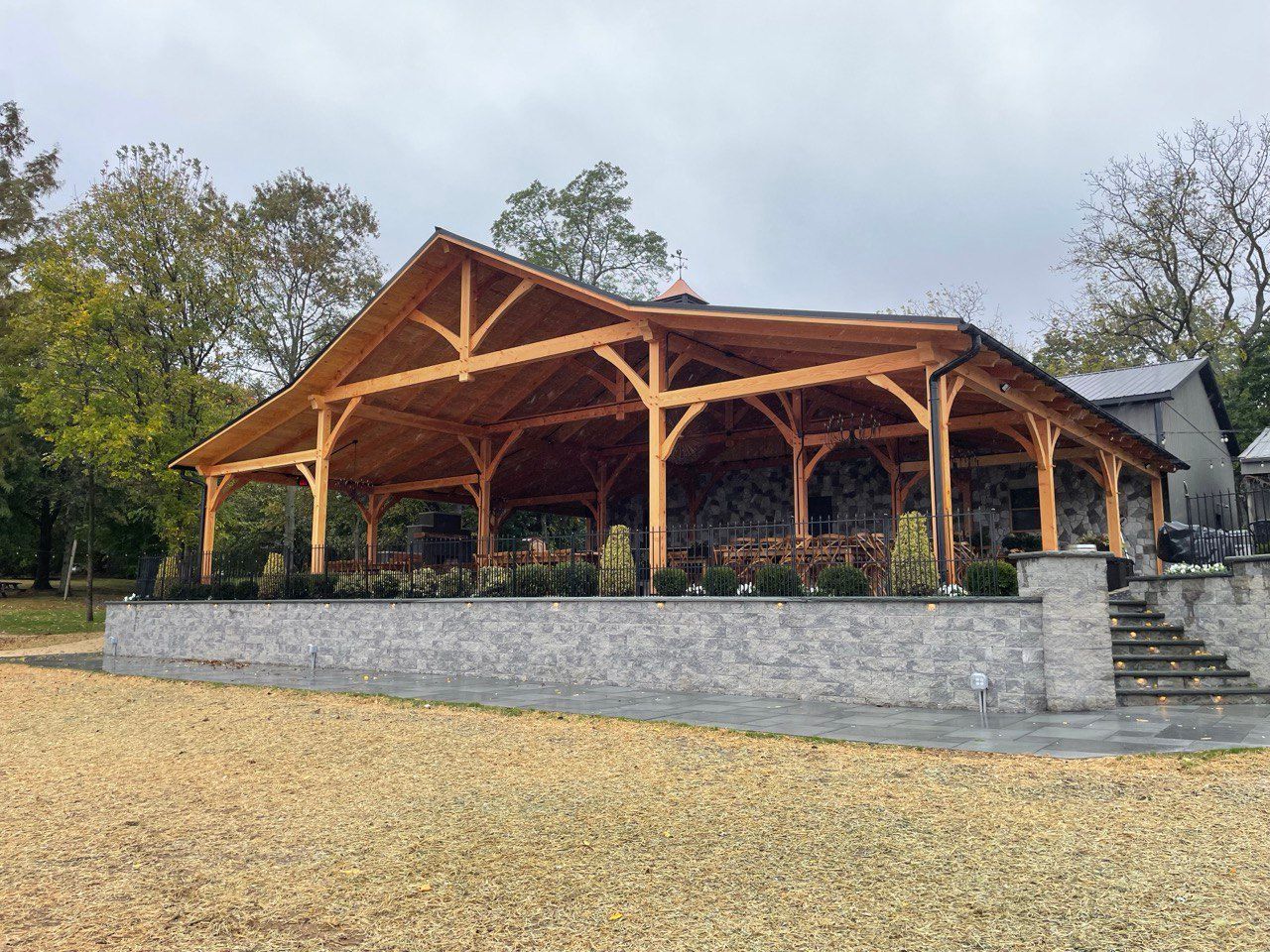 Wooden-framed pavilion with stone base and gravel ground; outdoor setting with trees under a cloudy sky.