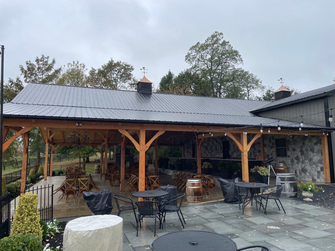 Outdoor pavilion with wooden beams, tables, and a dark metal roof. Cloudy sky.