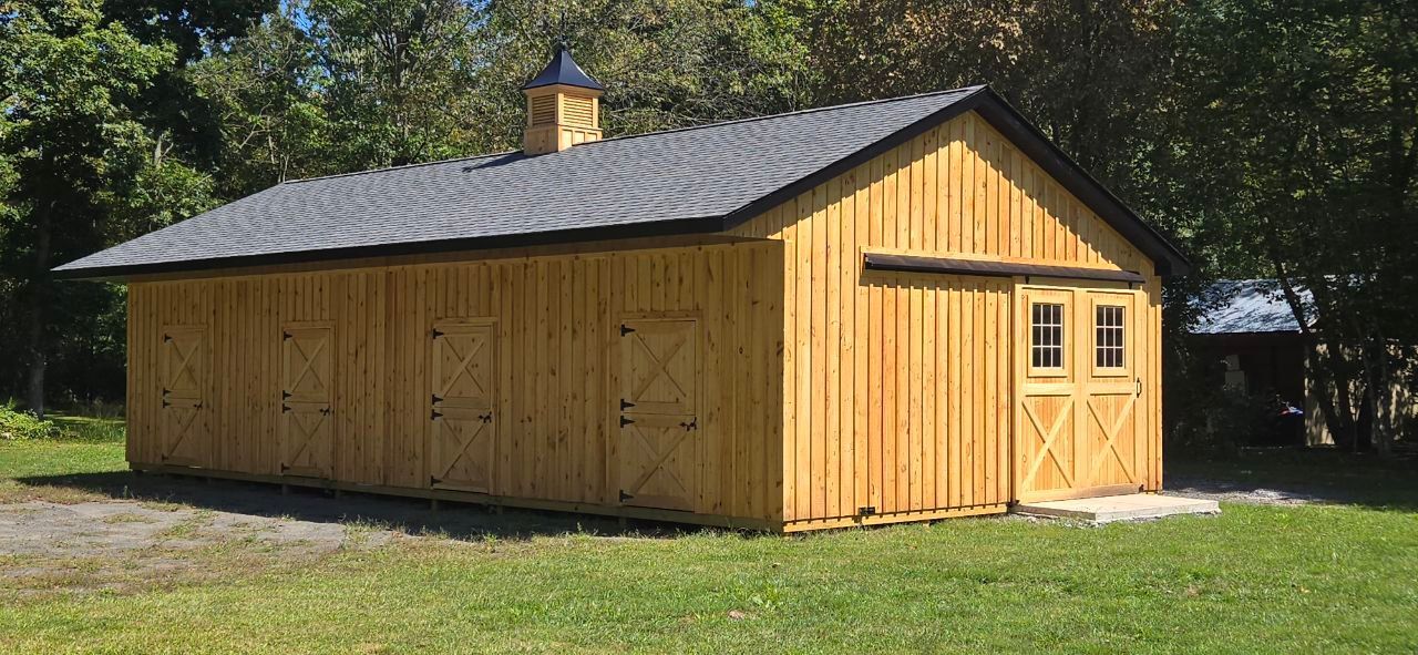 Yellow wooden barn with gray roof and cupola, set on a grassy lawn with trees in the background.
