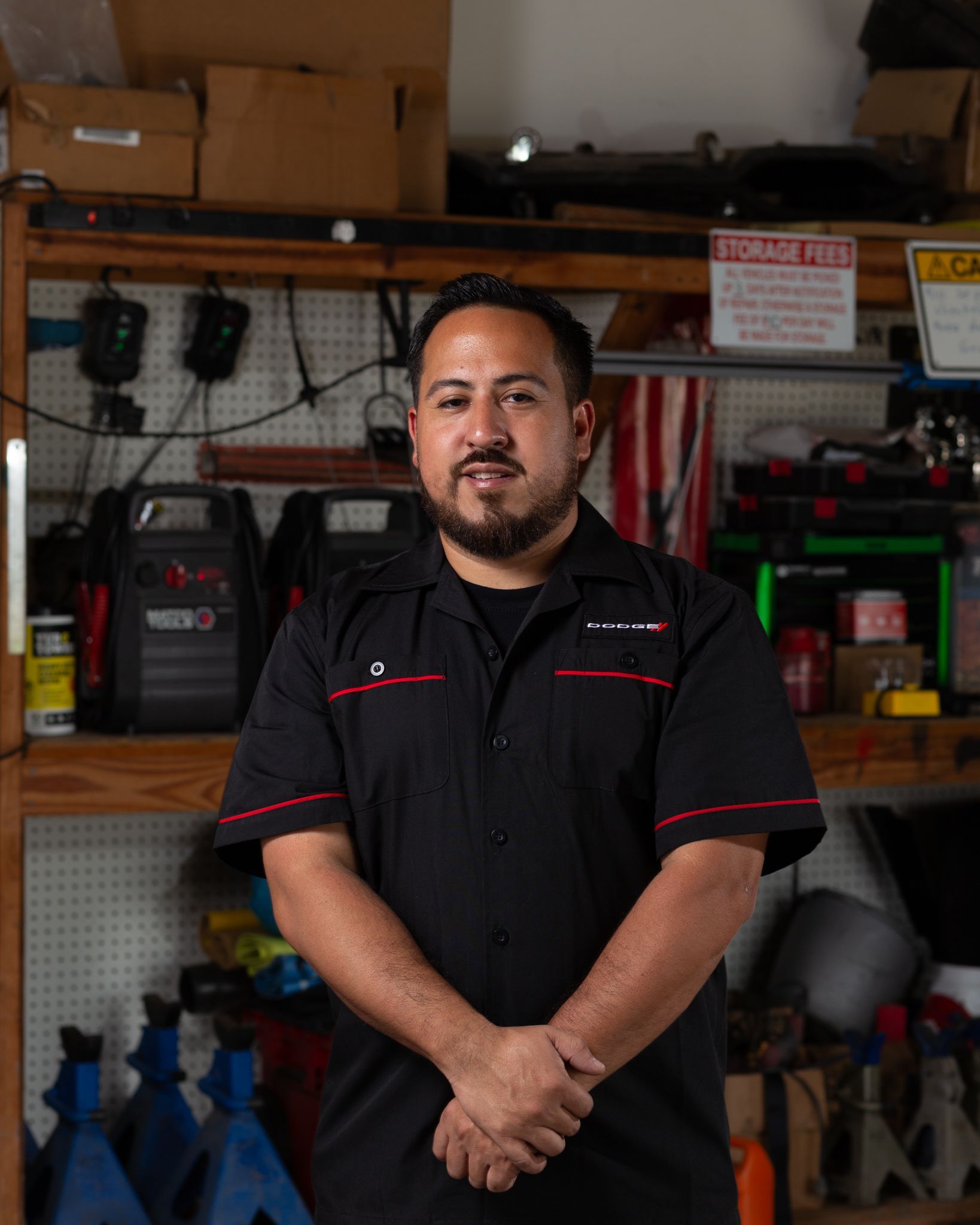 Mechanic in a black shirt, arms crossed, standing in a cluttered garage, smiling slightly.