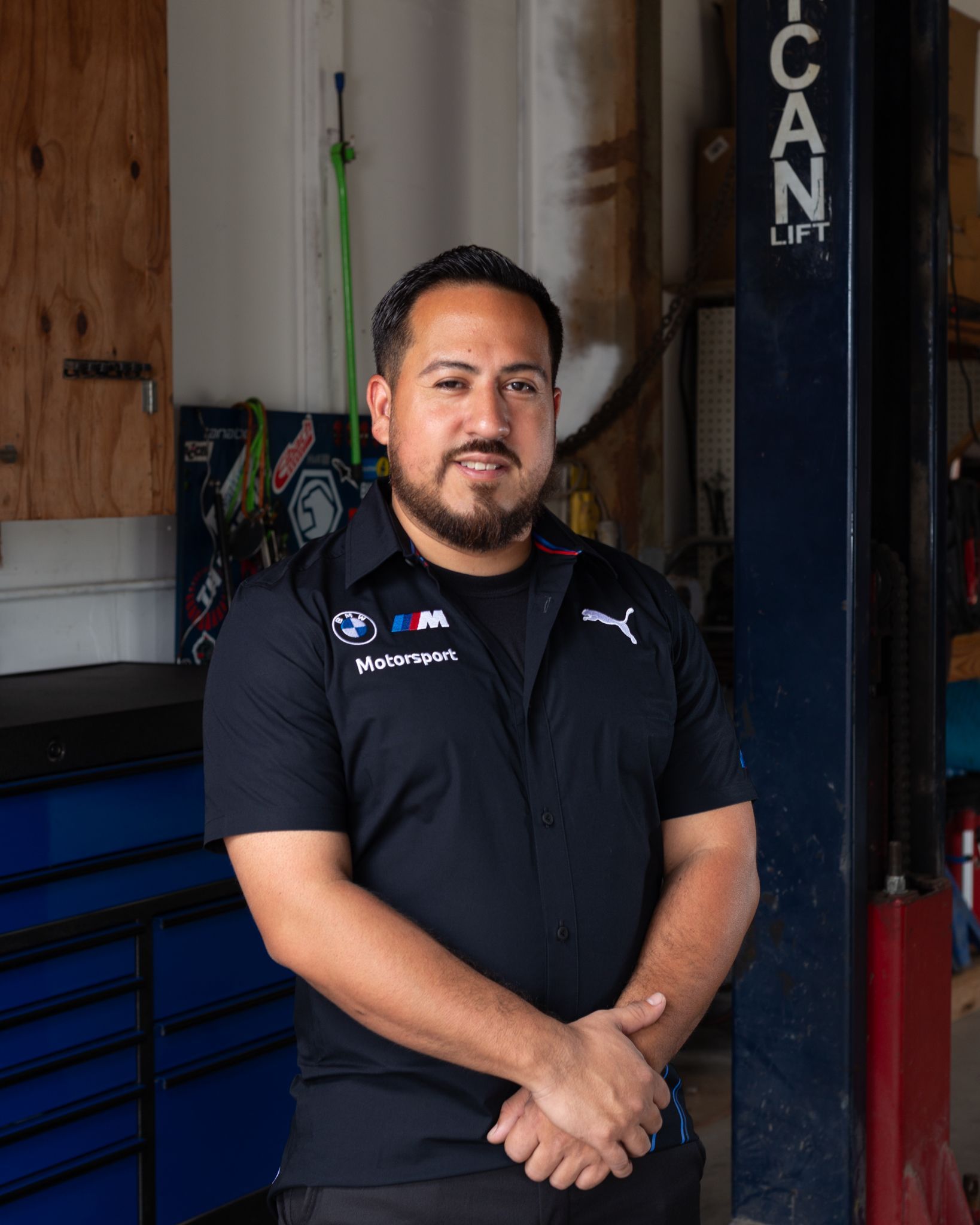 Man in black mechanic uniform with arms crossed in a garage setting, near a car lift.
