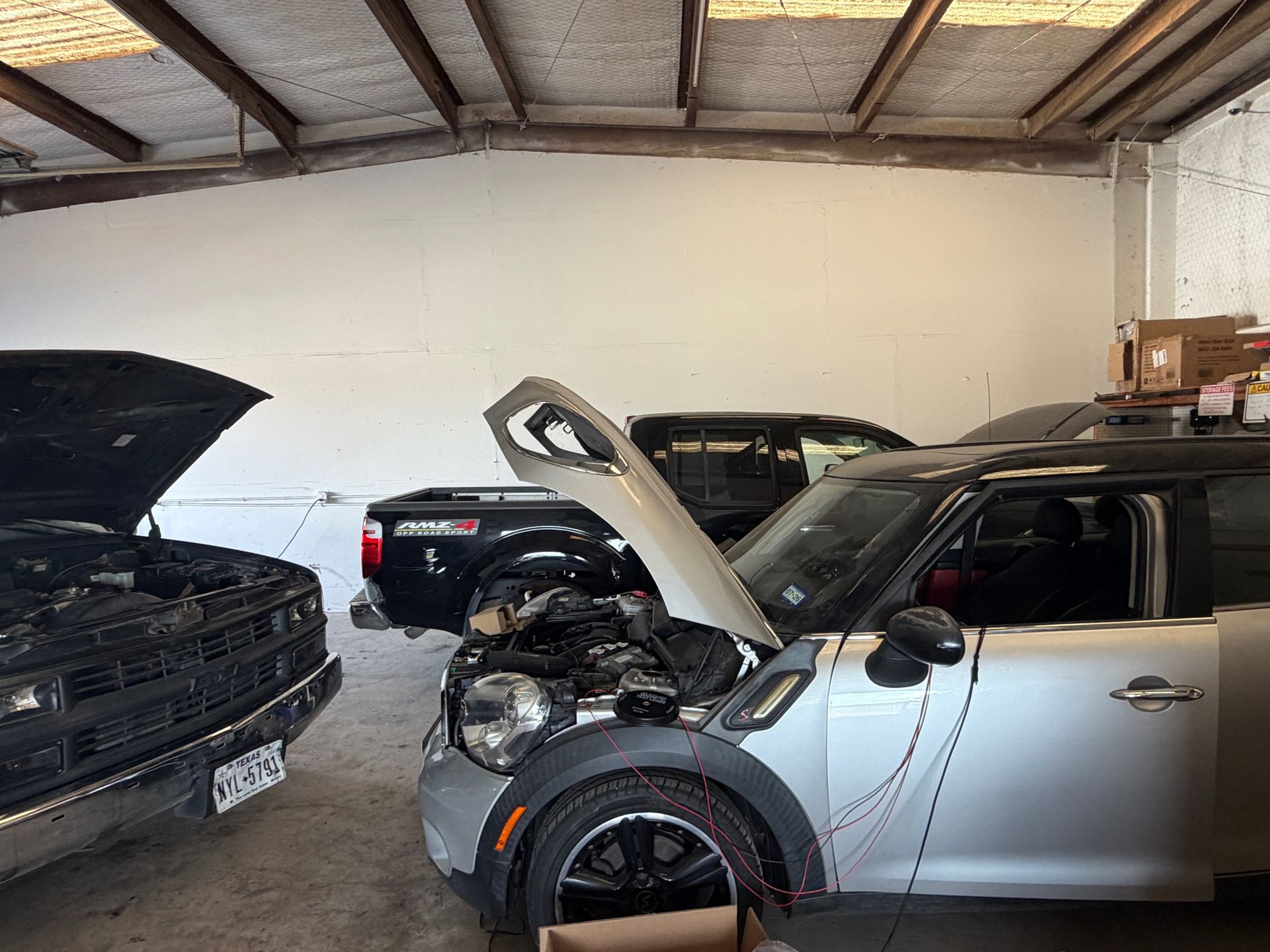 Cars in a garage with open hoods. Silver Mini Cooper in the foreground, black truck behind, and a black older truck on the left.
