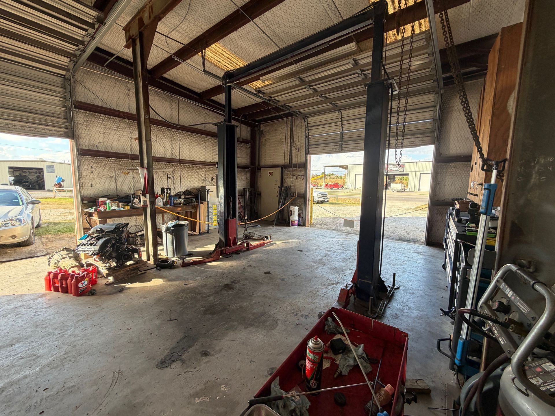 Interior of a car repair shop with two-post lift; tools, parts, and open bay doors are visible.