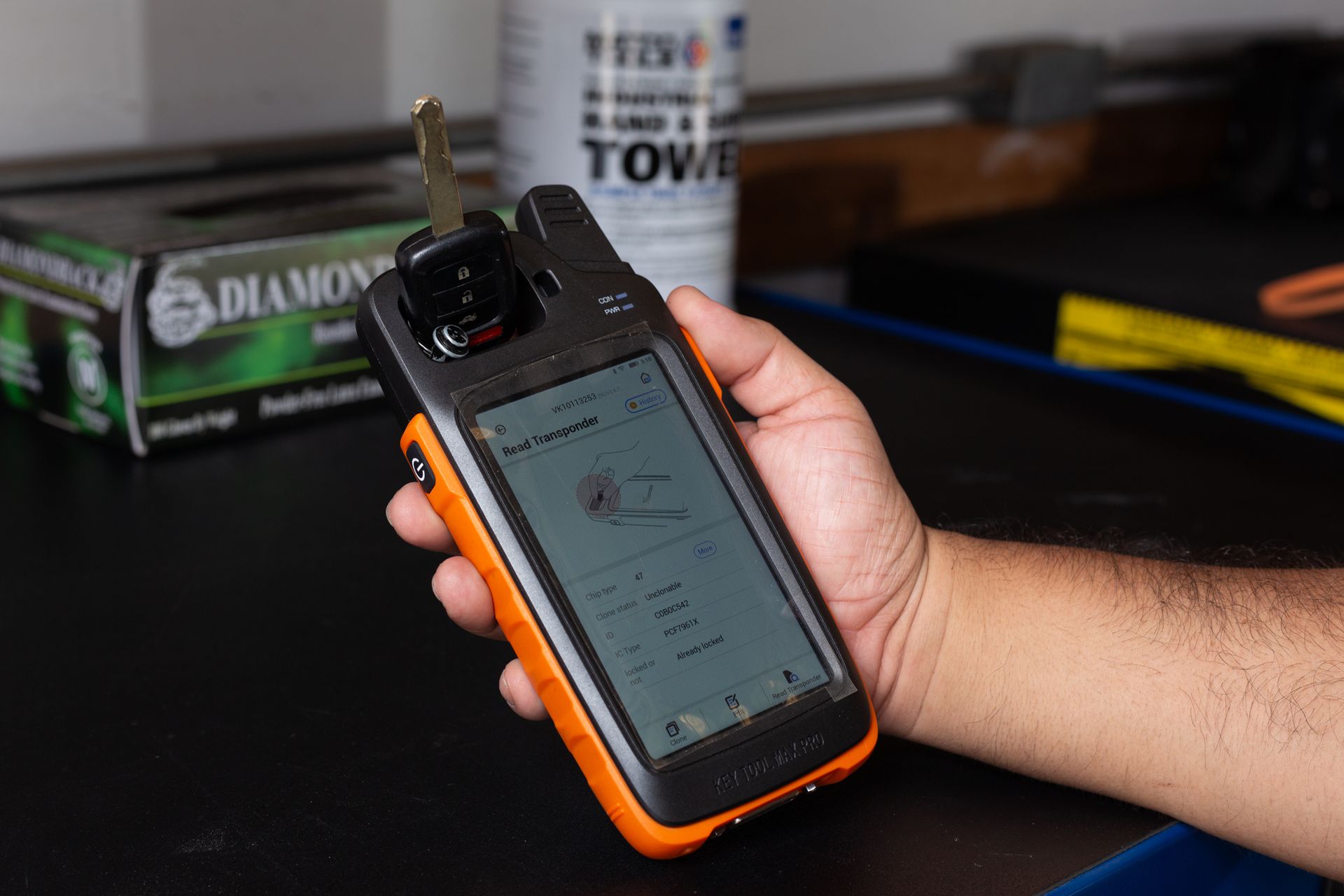 Hand holding a key programming device with a key attached, black and orange color scheme, indoors.