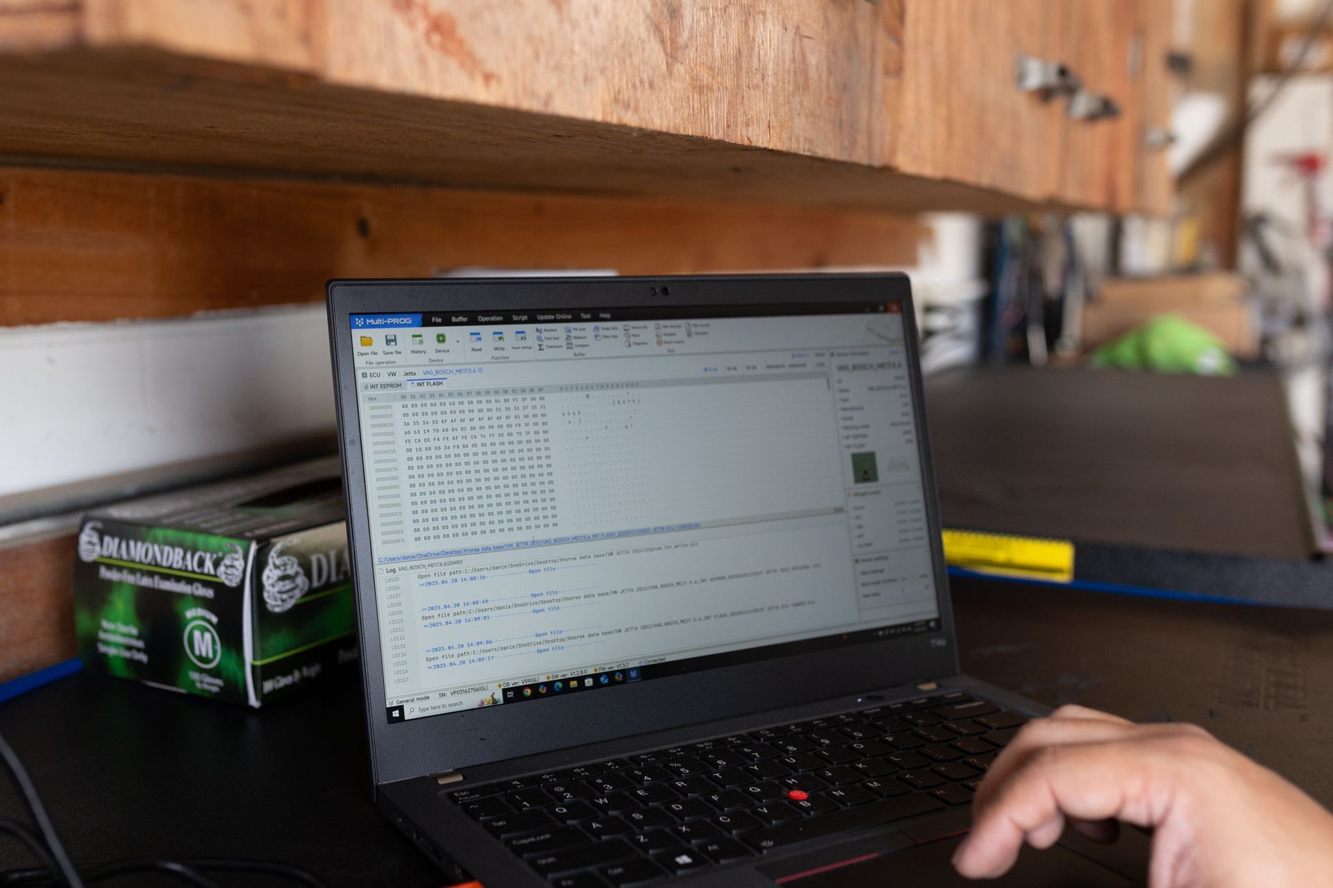 Person working on laptop in a cluttered workshop. Screen displays code, hand on trackpad.