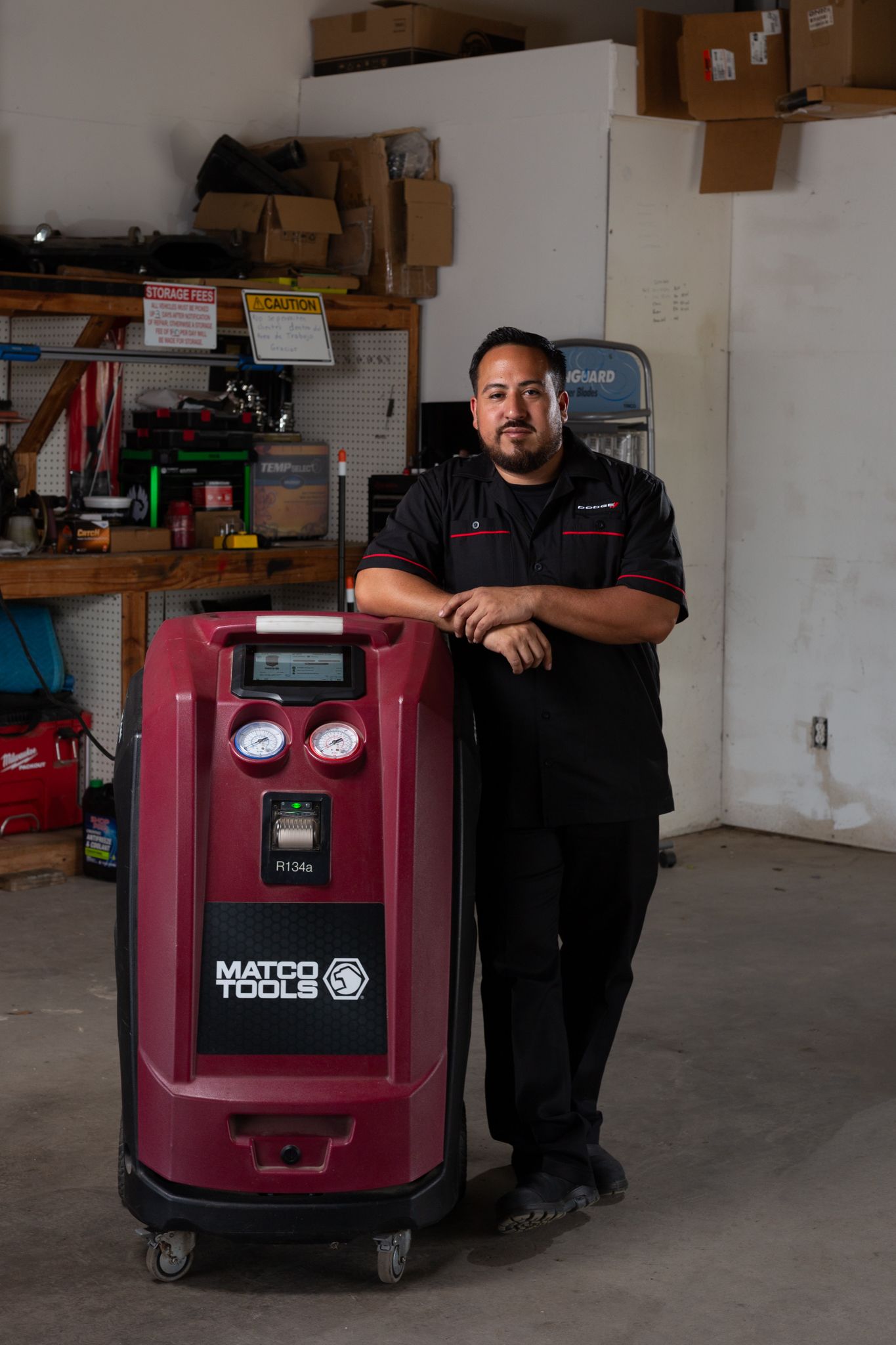 Mechanic in black uniform leans on an automotive air conditioning machine in a shop.