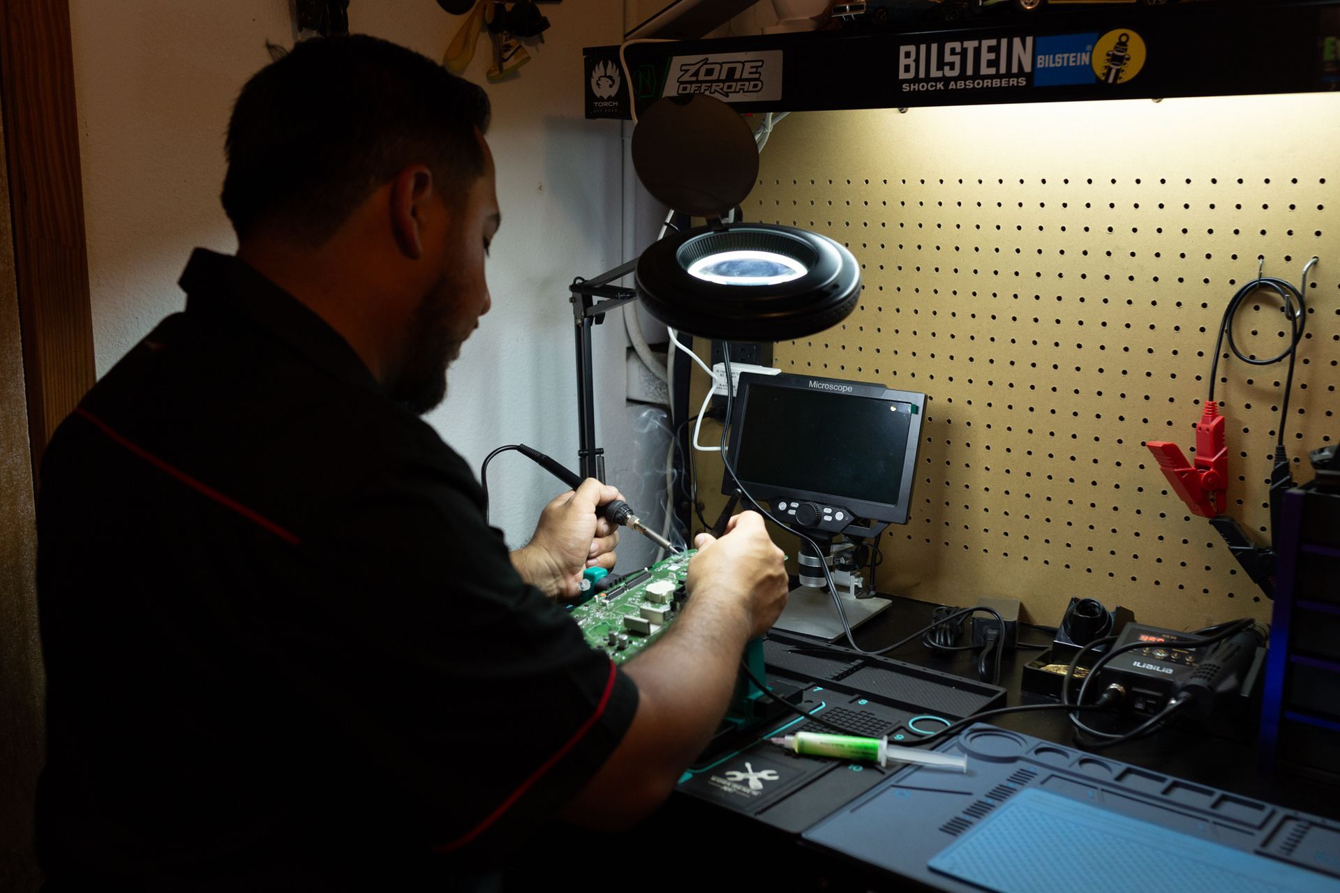 Man soldering circuit board at a workbench, with magnifying lamp and monitor. Workshop setting.