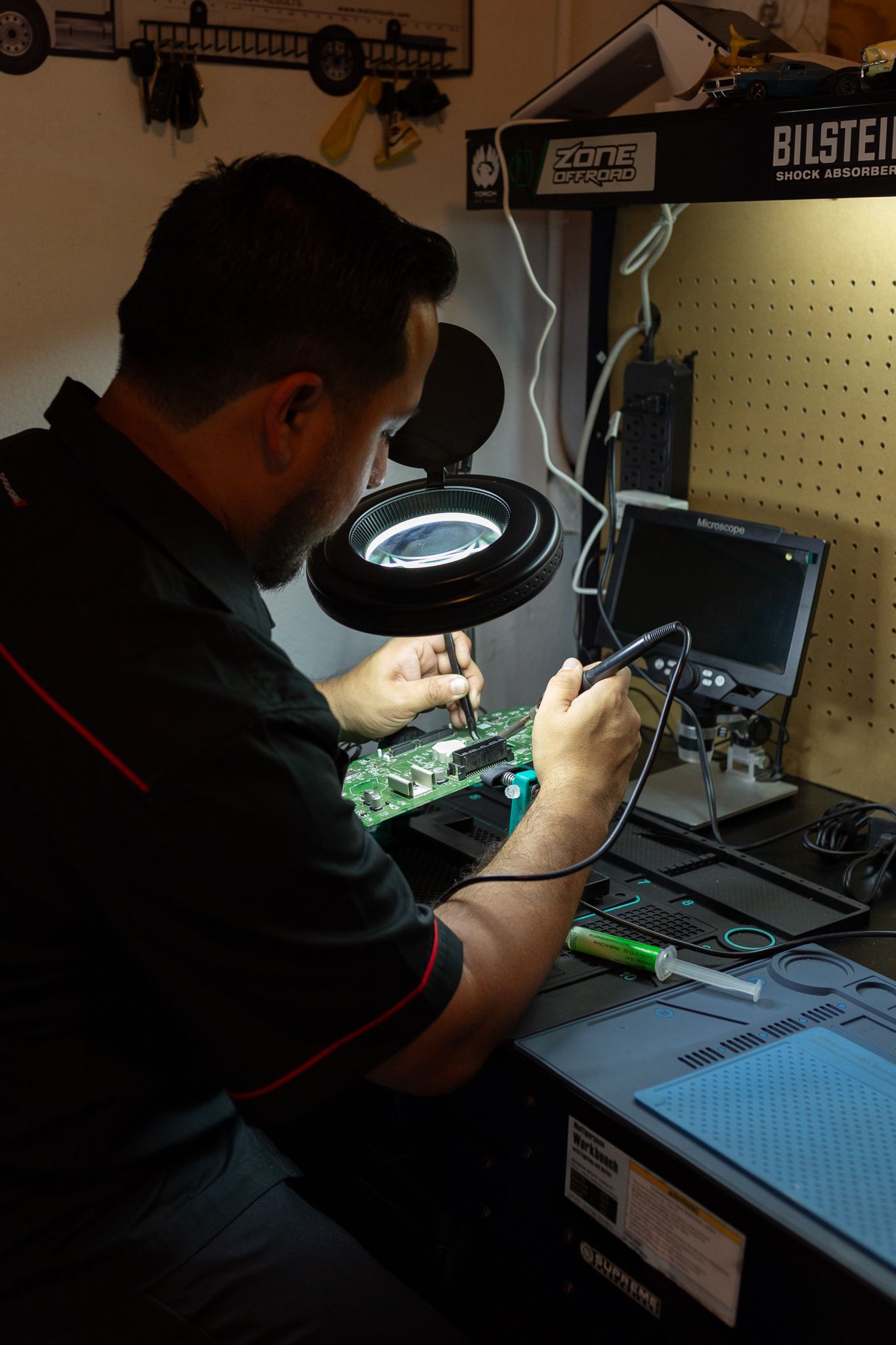 Man soldering electronic circuit board, using a magnifying glass and a laptop at a workbench.