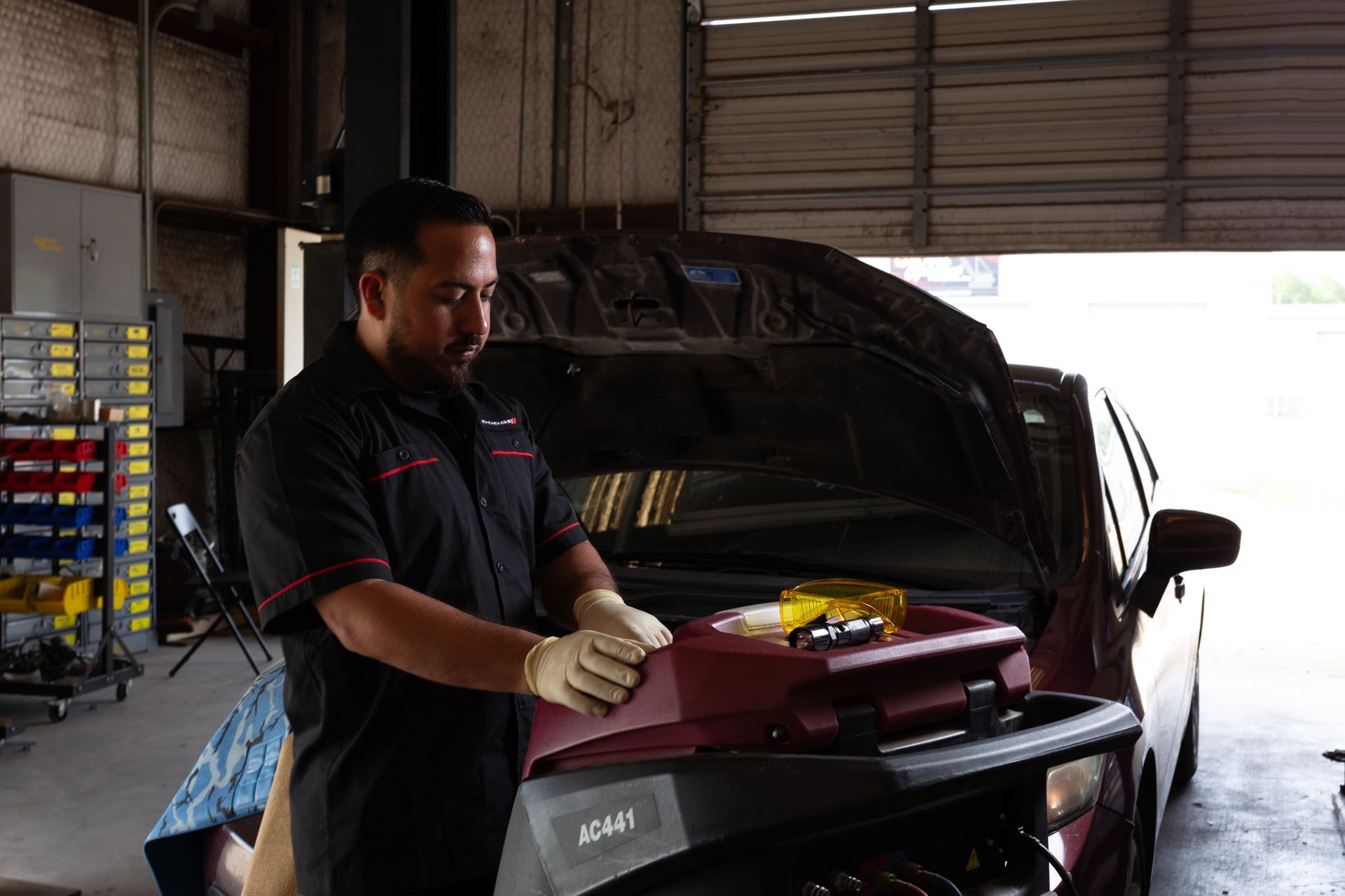 Mechanic working on a car in a shop, looking under the hood. He wears gloves and a dark uniform.