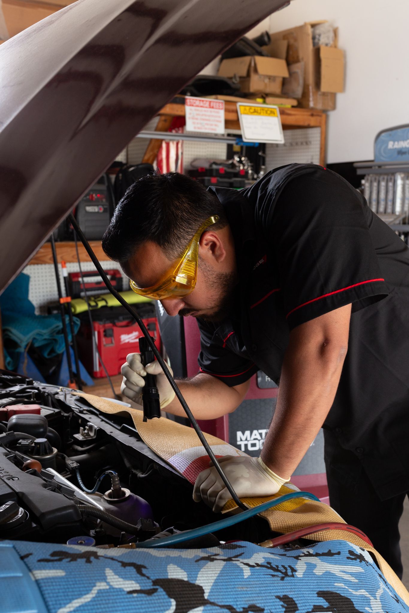 Mechanic inspects car engine, wearing safety glasses and gloves, in a garage.