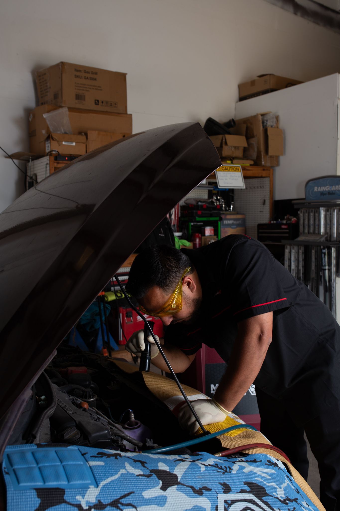 Mechanic working on a car engine with the hood up, wearing safety glasses, in a garage.