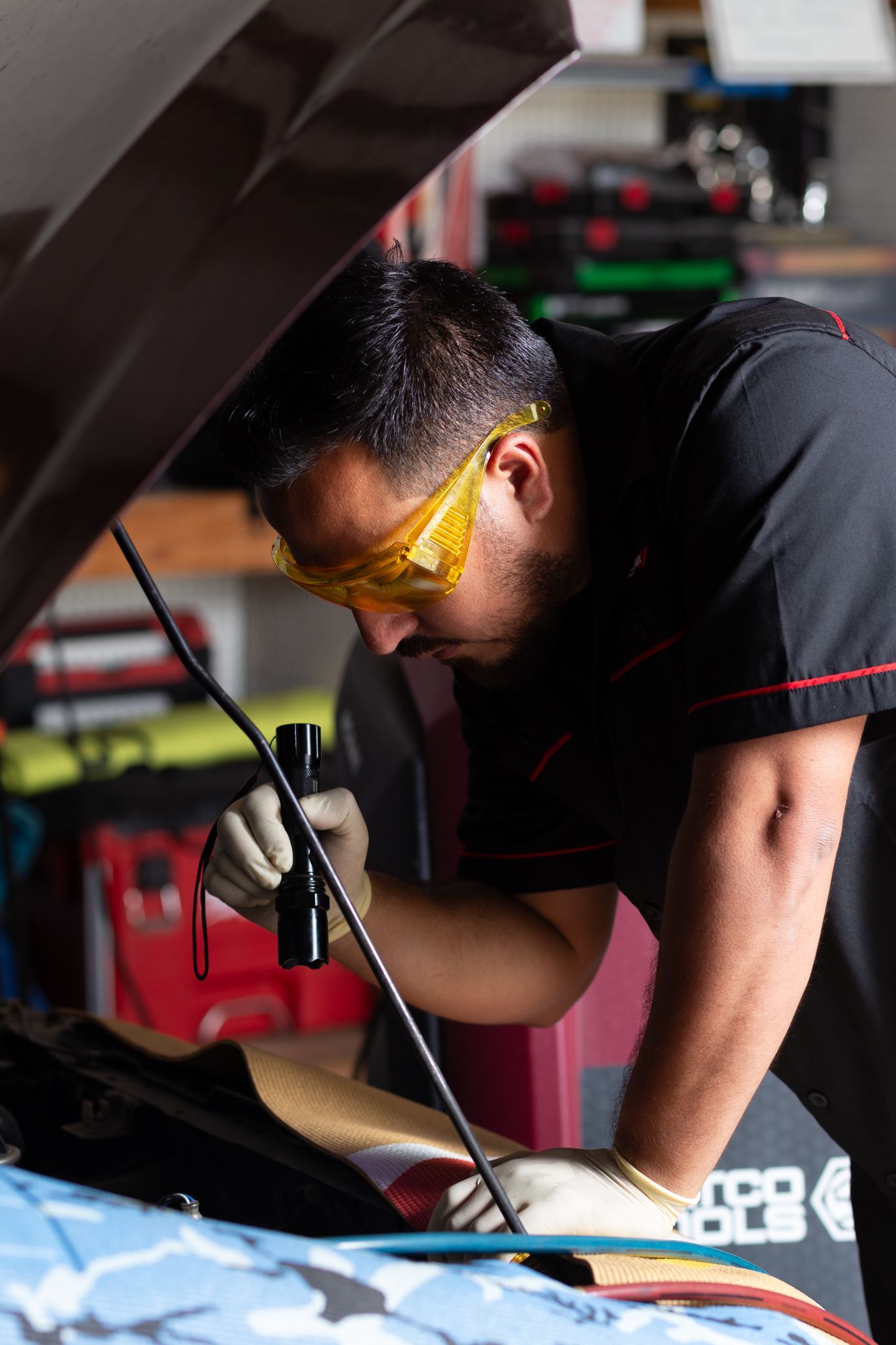 Mechanic using a flashlight to inspect an engine, wearing safety glasses and gloves, at a car repair shop.
