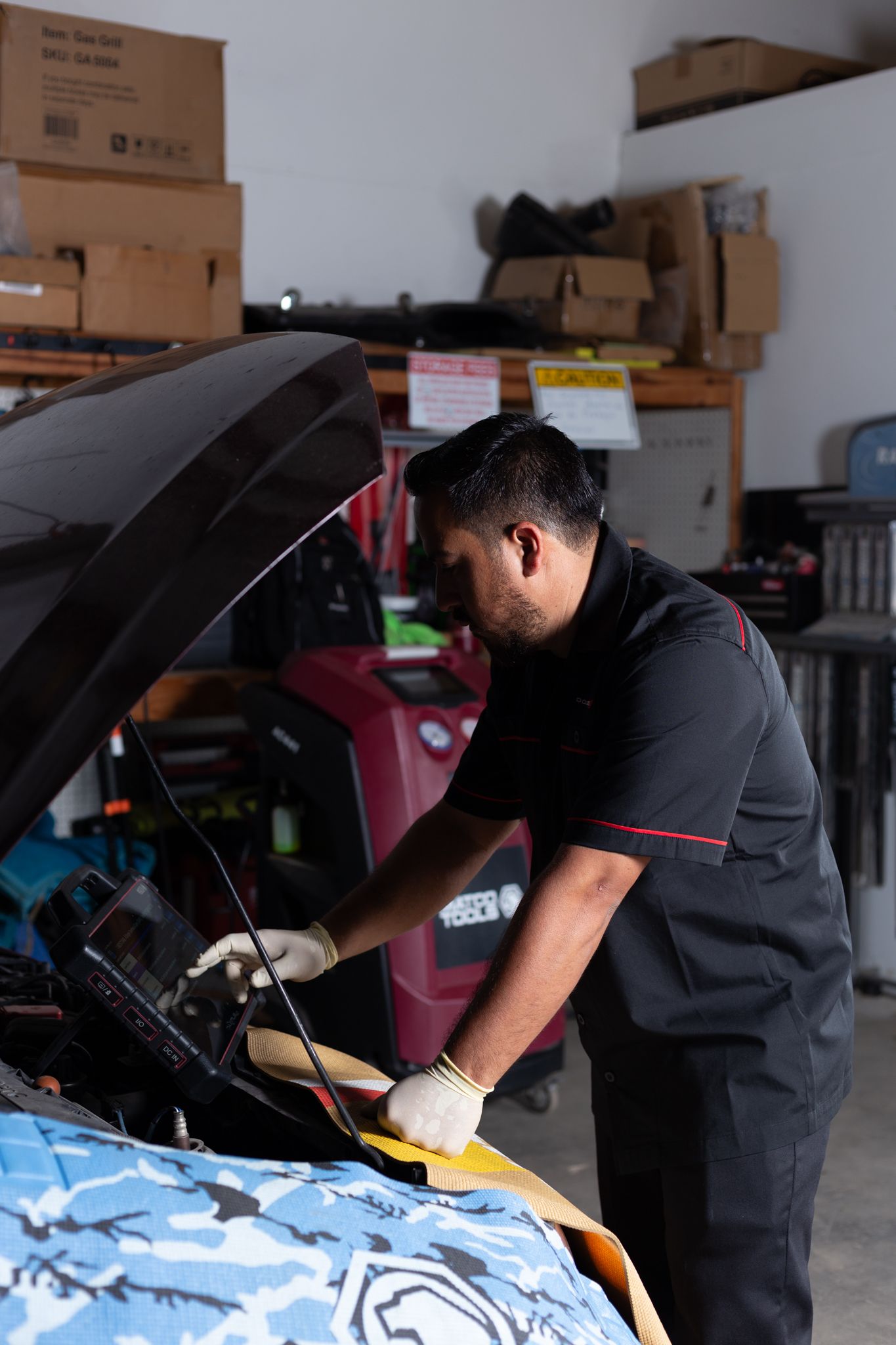 Mechanic working on a car with the hood up, using a diagnostic tool in a garage.
