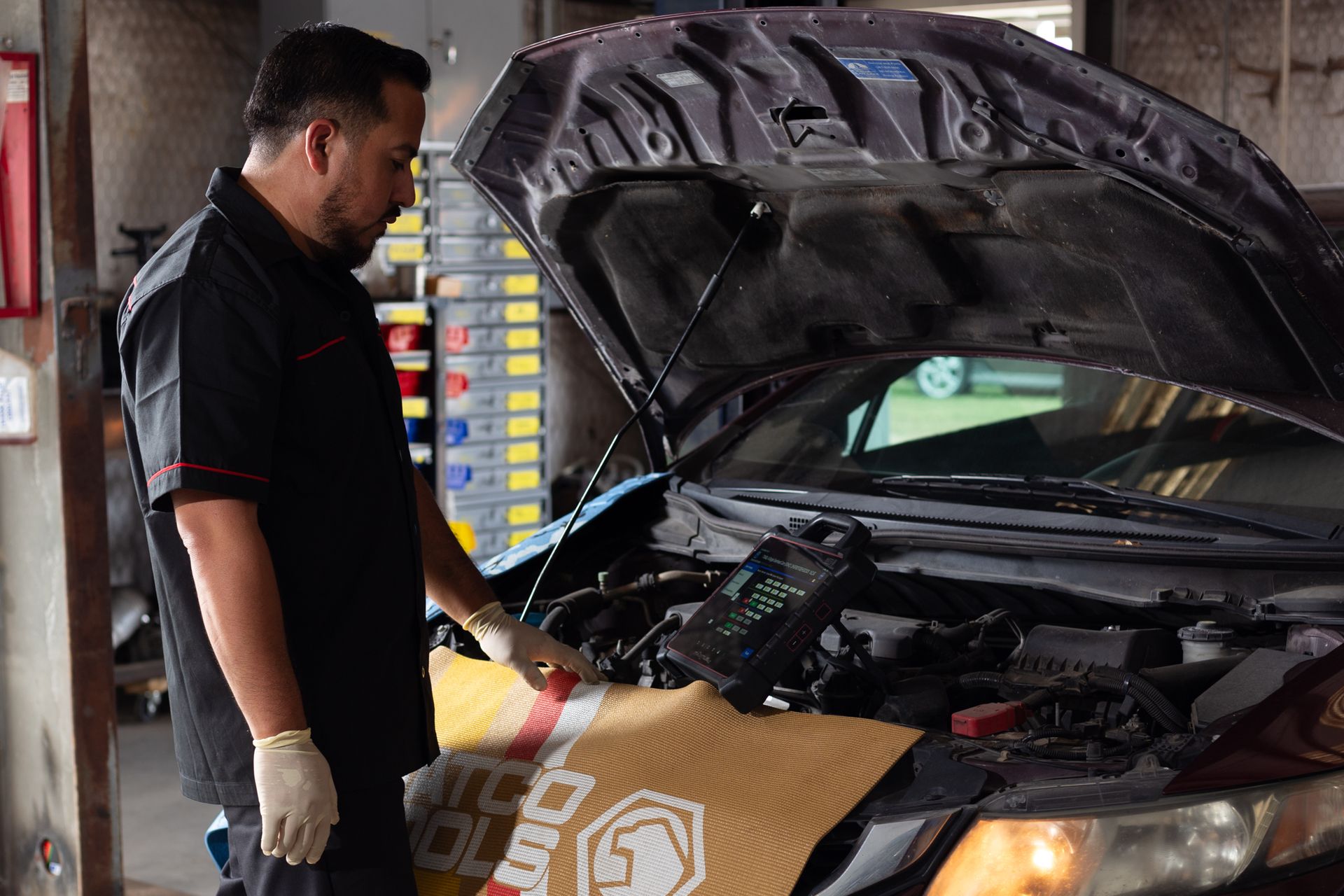 Mechanic working on car engine, using diagnostic tool. Garage setting, open hood, black car, wearing gloves.