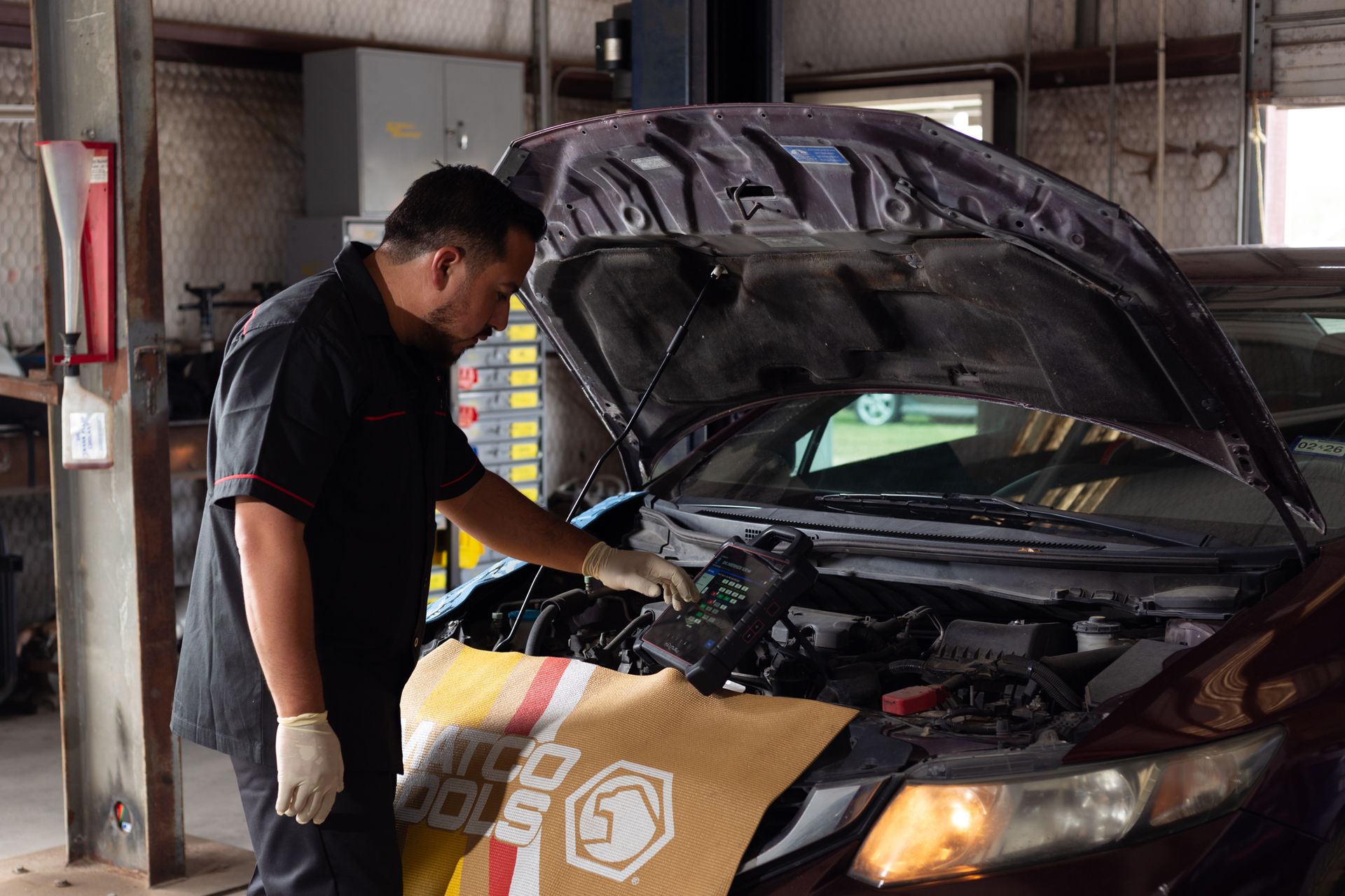 Mechanic using a diagnostic tool on a car with its hood open in a garage.